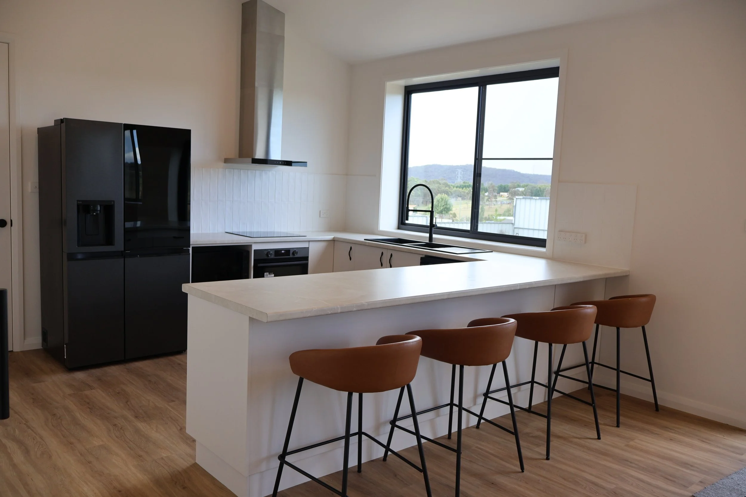 Modern kitchen with black refrigerator, white cabinets, large window with scenic view, kitchen island with four brown chairs, and wood flooring.