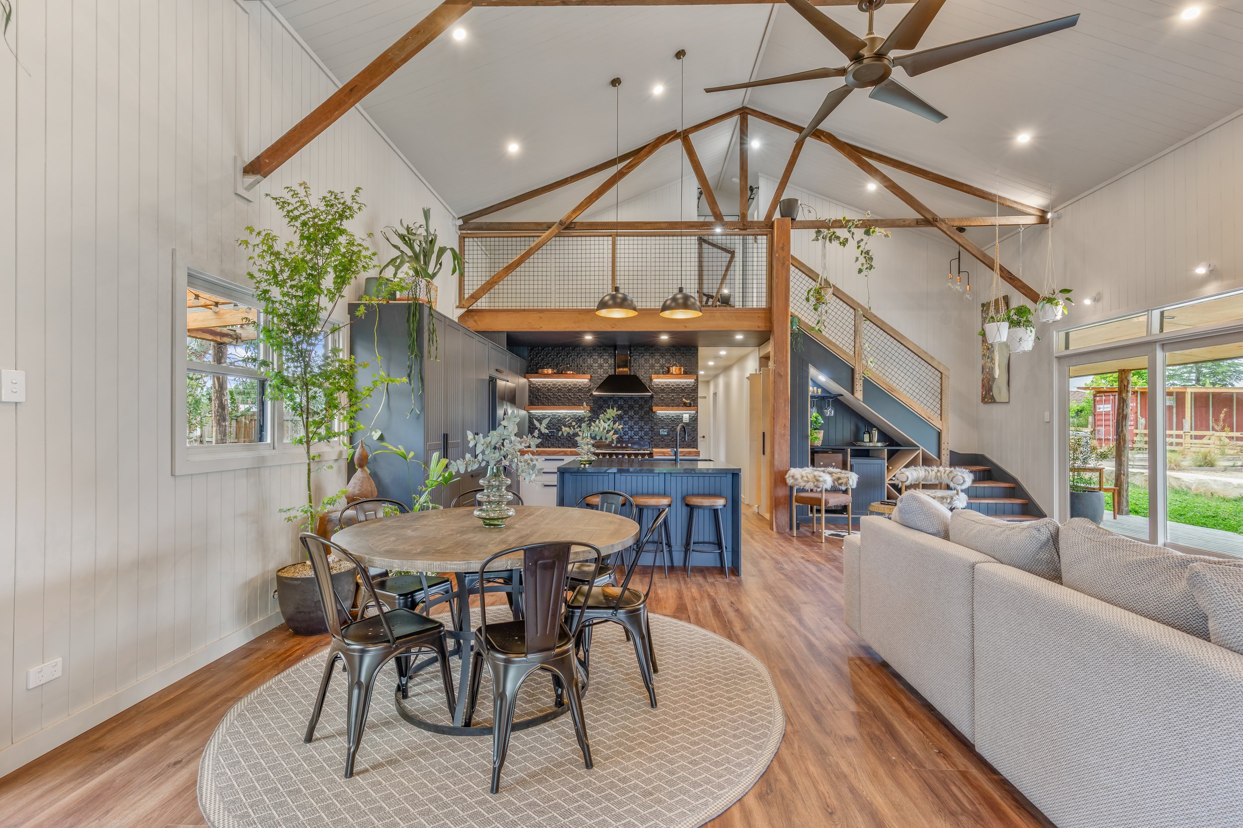 Interior view of a modern open-concept living space with a kitchen, dining area, and sitting area. The high vaulted ceiling features exposed wooden beams and a ceiling fan. The room has large windows and sliding doors, natural light, plants, and minimalist decor.