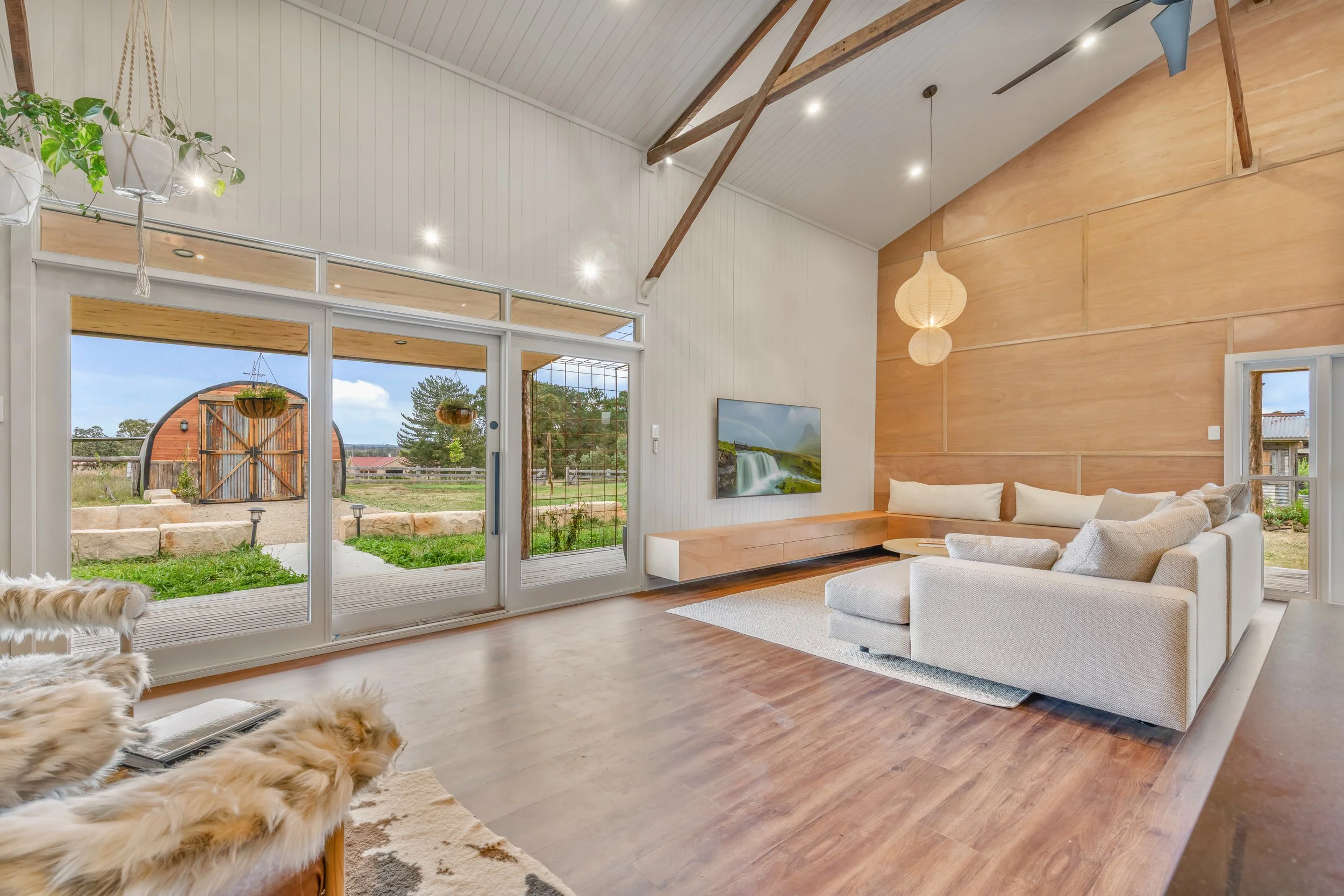 Modern living room with white and wooden walls, large sliding glass doors leading to an outdoor area, mounted TV, sectional sofa, and hanging paper lanterns, with a view of a barn and trees outside.