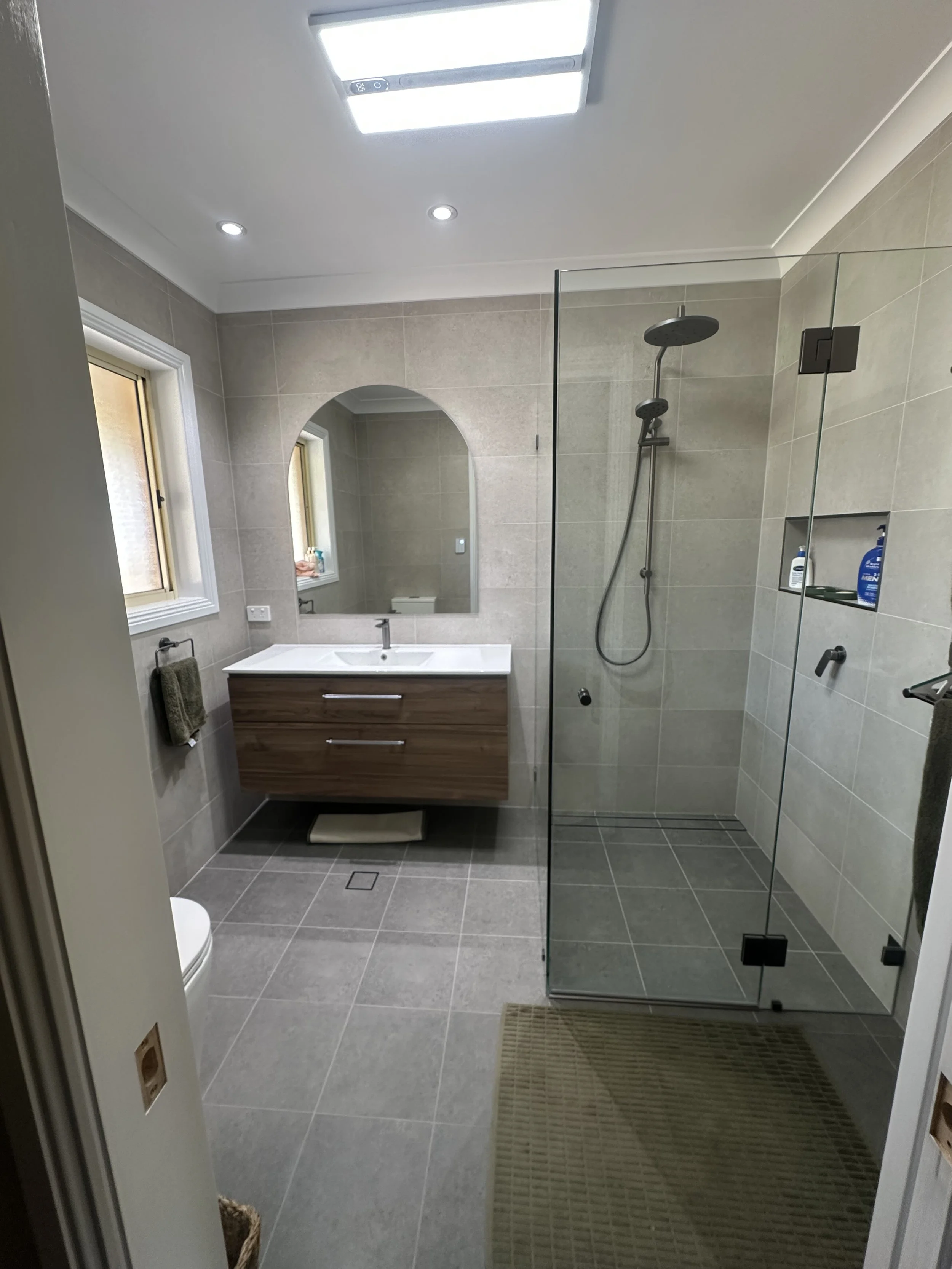 A modern bathroom with beige tiled walls and floor, a glass shower enclosure with a rainfall showerhead, a white sink vanity with a mirror, two small windows, a soap dispenser, and two towels hanging on a rack.
