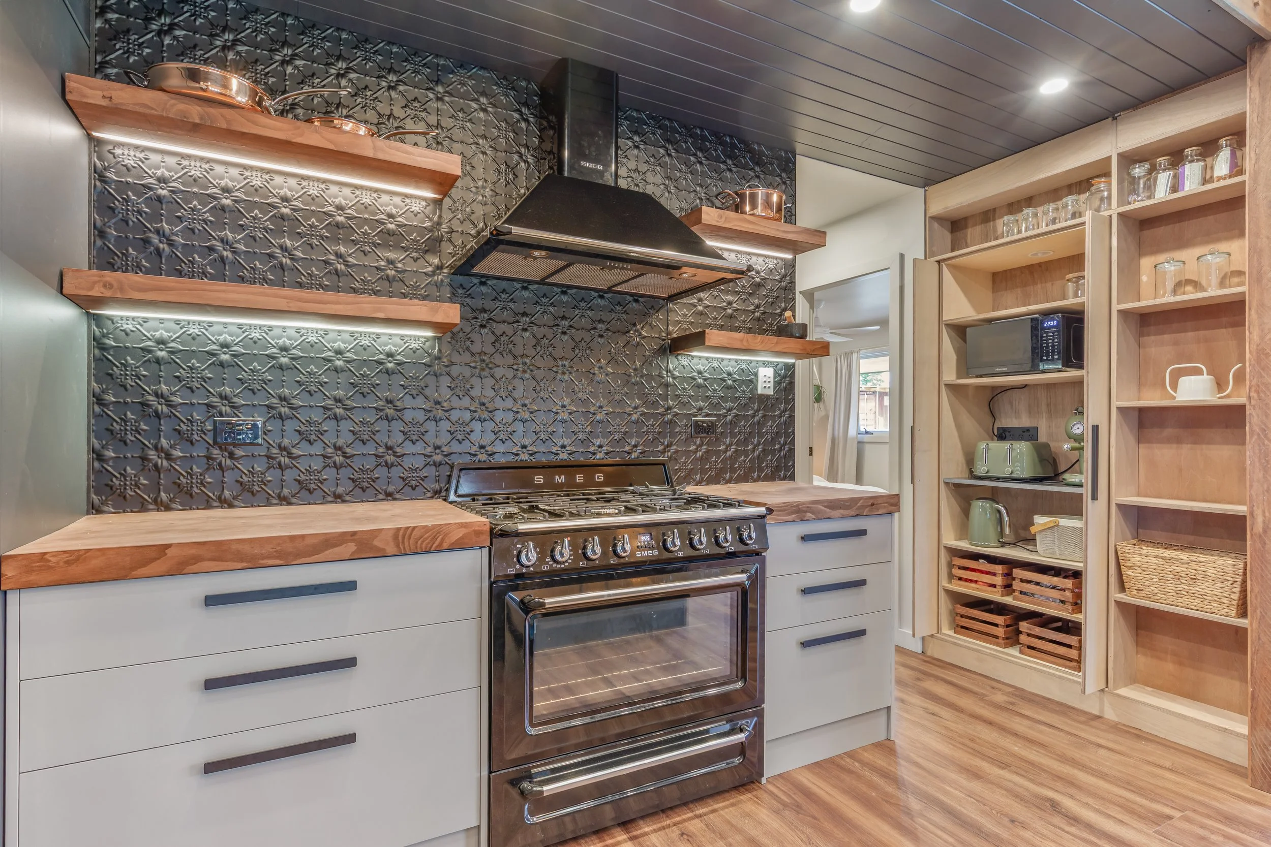 A modern kitchen with a black patterned tiled backsplash, a stainless steel oven, wooden shelves with copper pots, a microwave, and wooden storage baskets.