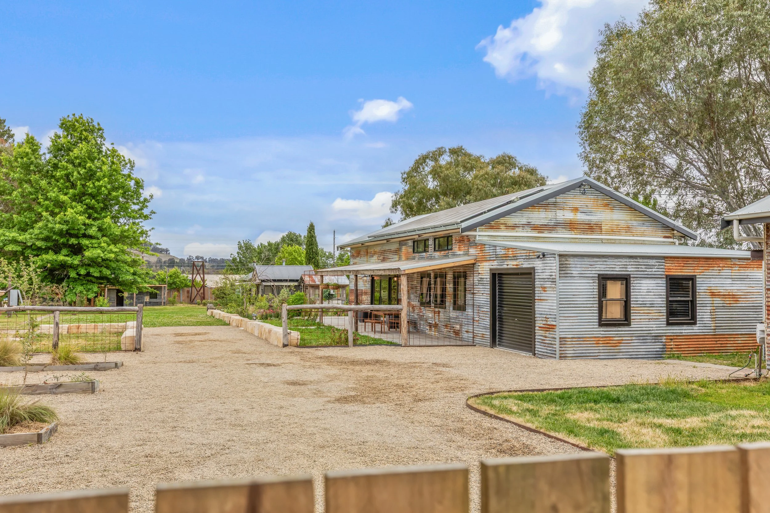 A rustic house in a rural setting with a gravel yard, surrounded by green trees and a wooden fence, under a partly cloudy sky.