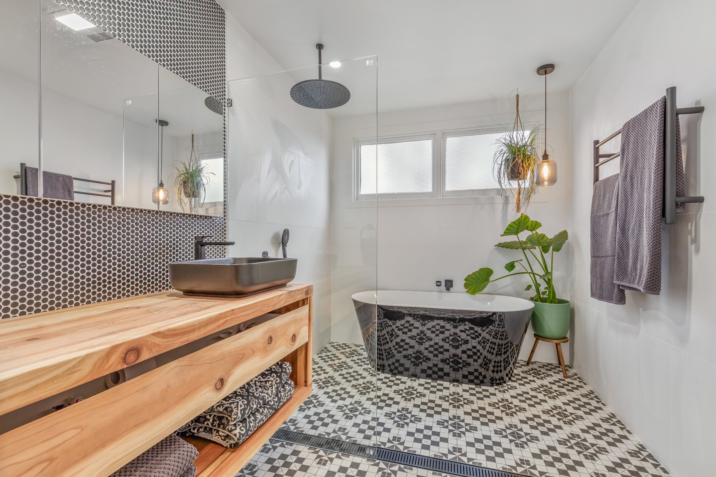 Modern bathroom featuring a walk-in shower with a rain showerhead, a black freestanding bathtub, a wooden vanity with a black vessel sink, potted plants, and textured towel racks, with patterned black and white floor tiles and white walls.