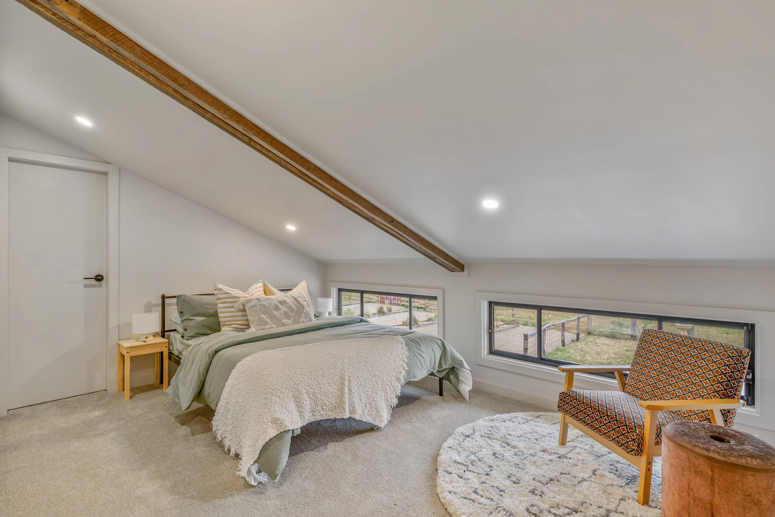 A cozy bedroom with a bed, side table, lamp, and a patterned armchair near a window showing an outdoor yard.