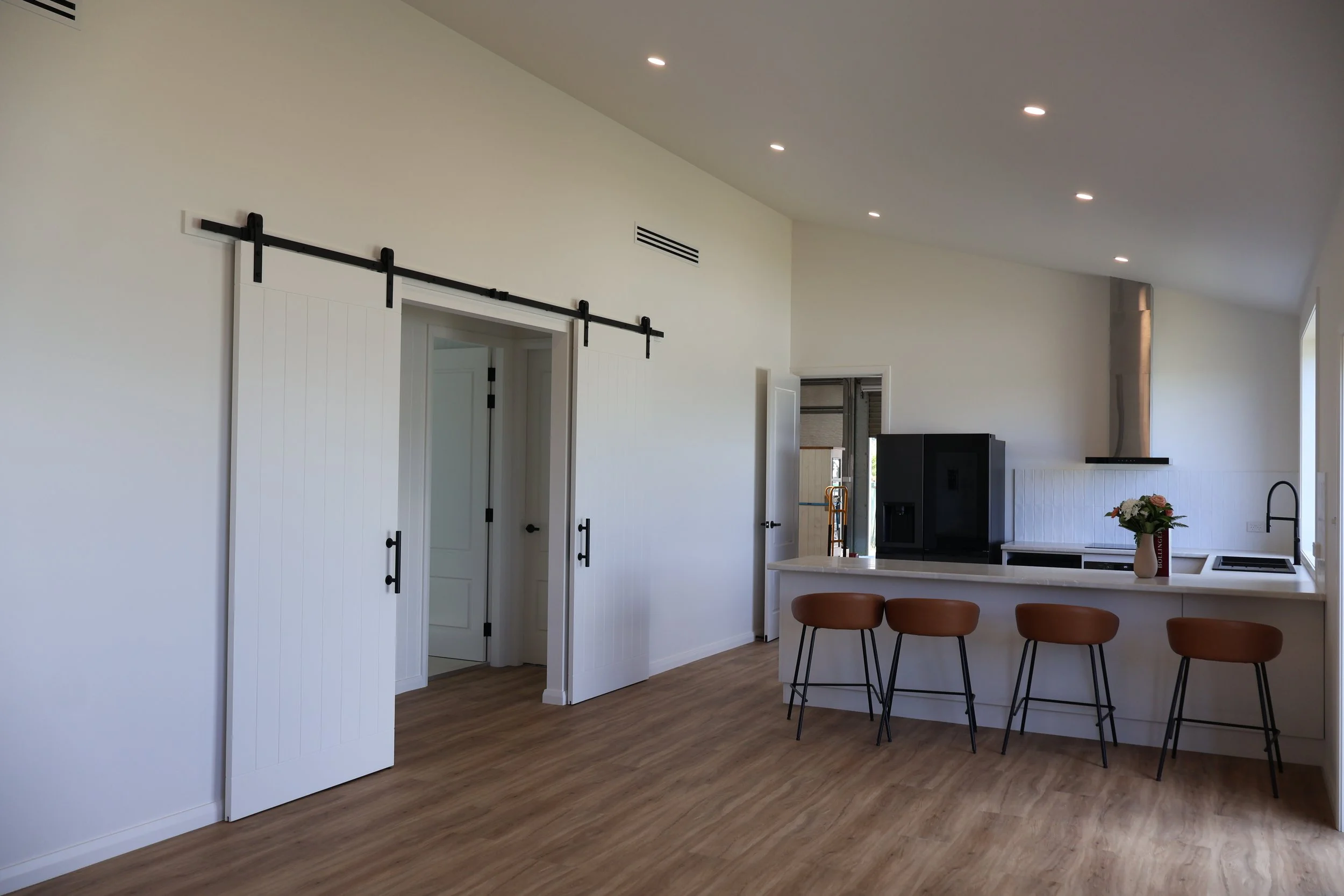 Modern open-concept kitchen and dining area with white walls, wood flooring, and ceiling lights. There is a kitchen island with a flower arrangement, and a set of brown barstools. Sliding barn door hardware is mounted on the wall, along with a visibl