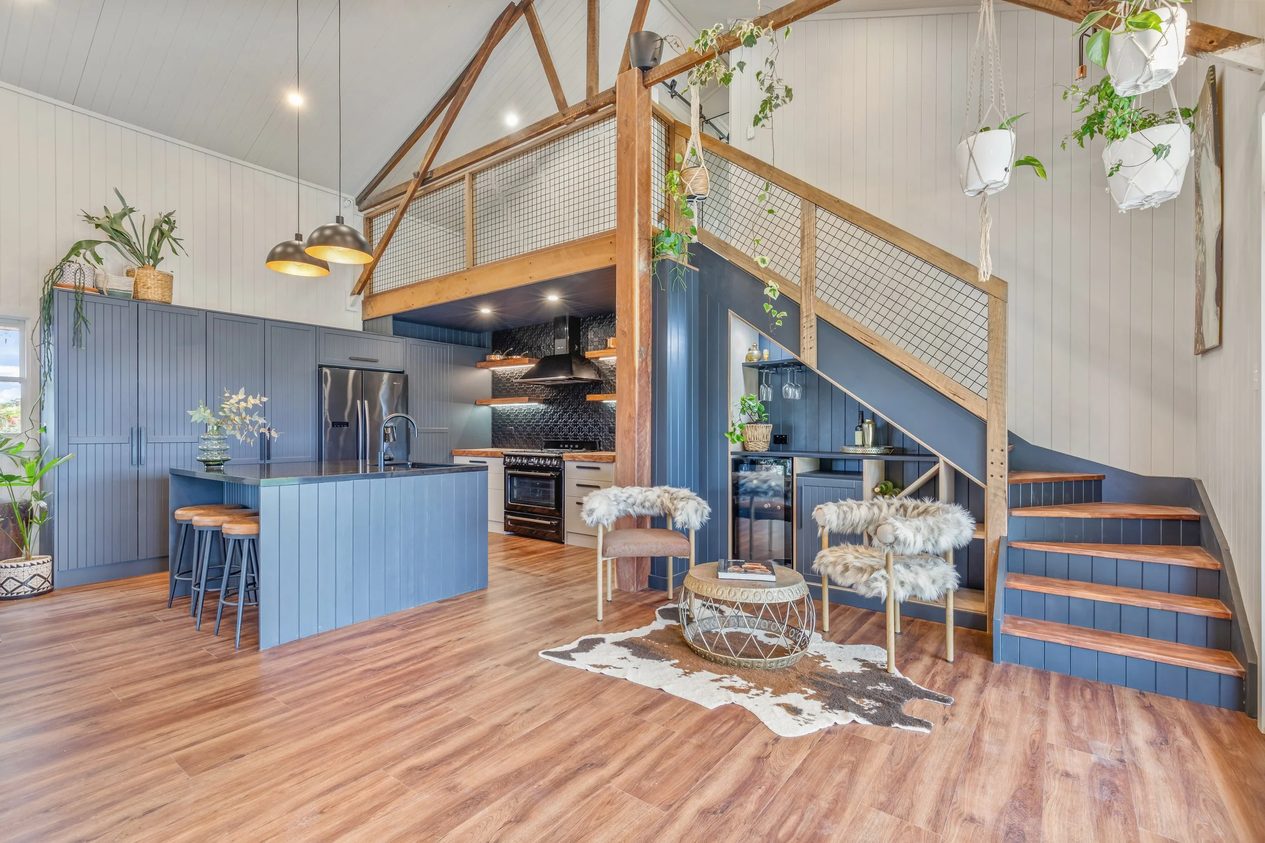 Open-concept kitchen and living room with high vaulted ceiling, wooden accents, blue cabinetry, and modern decor, including hanging planters and fur-covered chairs.