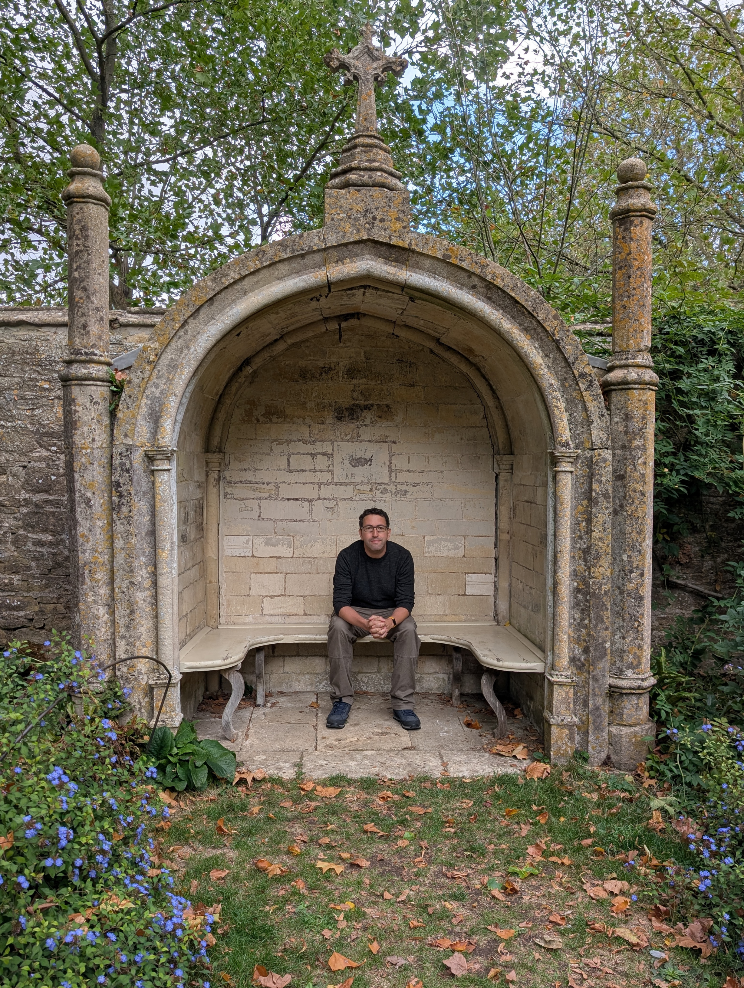 Man sitting on a stone bench with an arched stone shelter behind him, surrounded by greenery and fallen leaves.