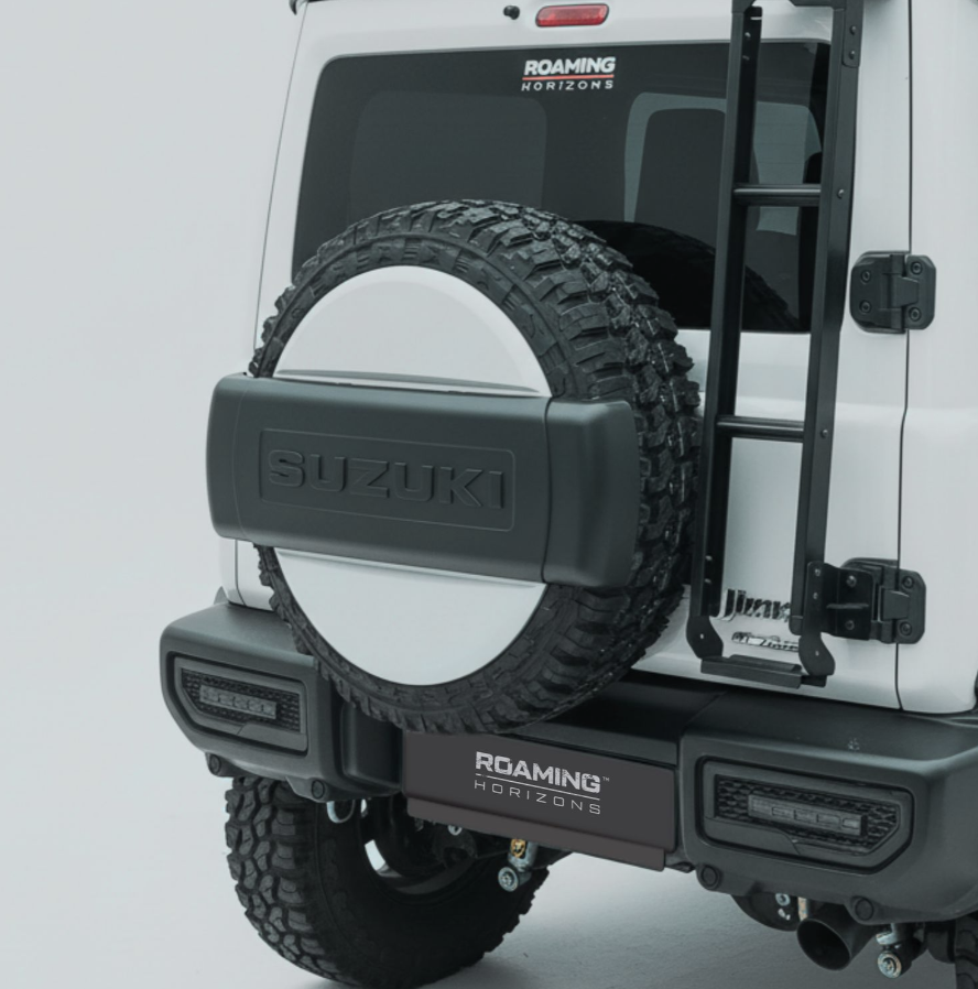 Rear view of a white Suzuki off-road vehicle with a spare tire mounted on the back, a ladder on the side, and various decals including 'Roaming Horizons'.