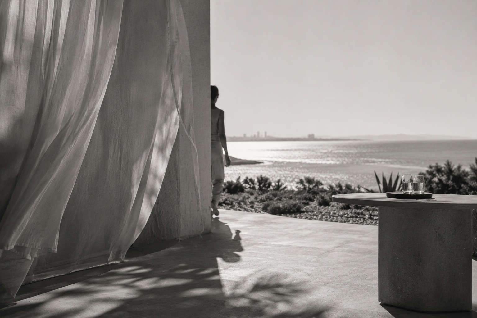 Black-and-white seaside terrace with flowing sheer curtains, a silhouetted figure, and a minimalist table overlooking the ocean.