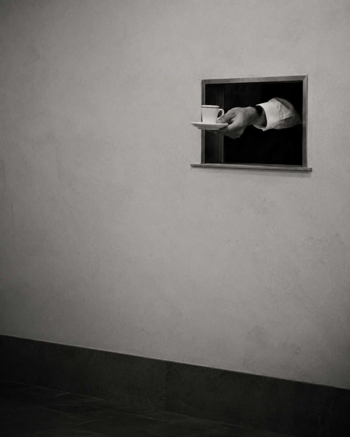 Black-and-white editorial image of a hand extending through a small service hatch, holding a tiny espresso cup and saucer against a minimalist wall.