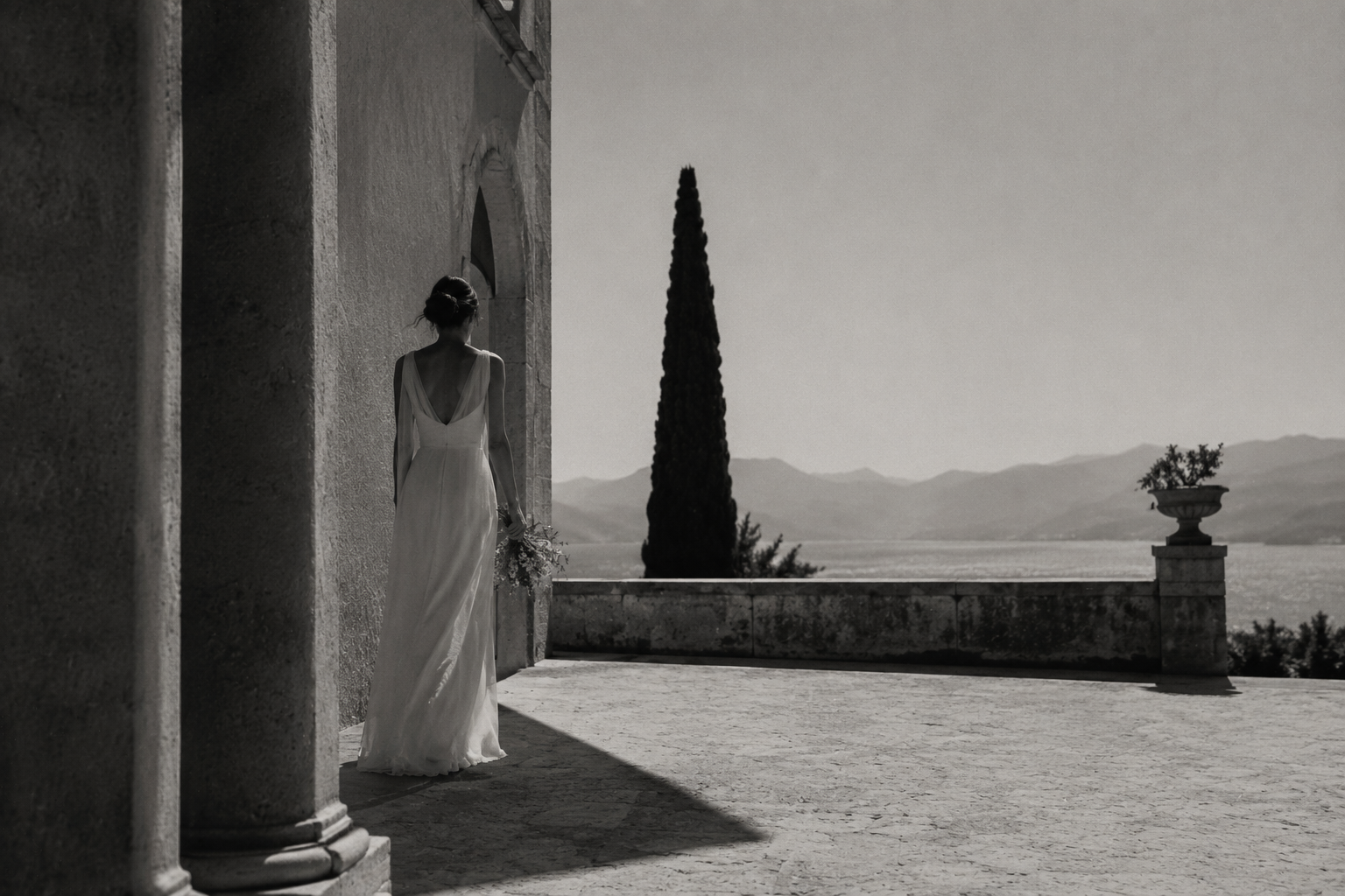 Black-and-white editorial image of a woman in a long white dress walking beside a historic stone building, with a tall cypress tree and distant mountains in the background.