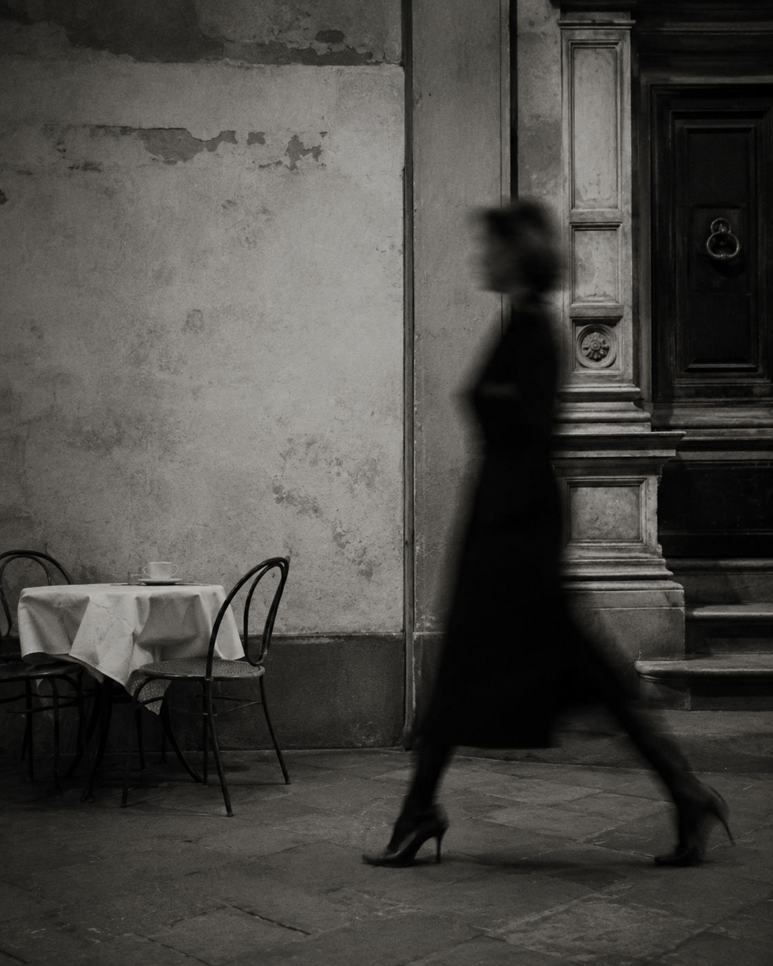 Black-and-white street scene with a blurred woman walking past a small café table and an ornate historic doorway.