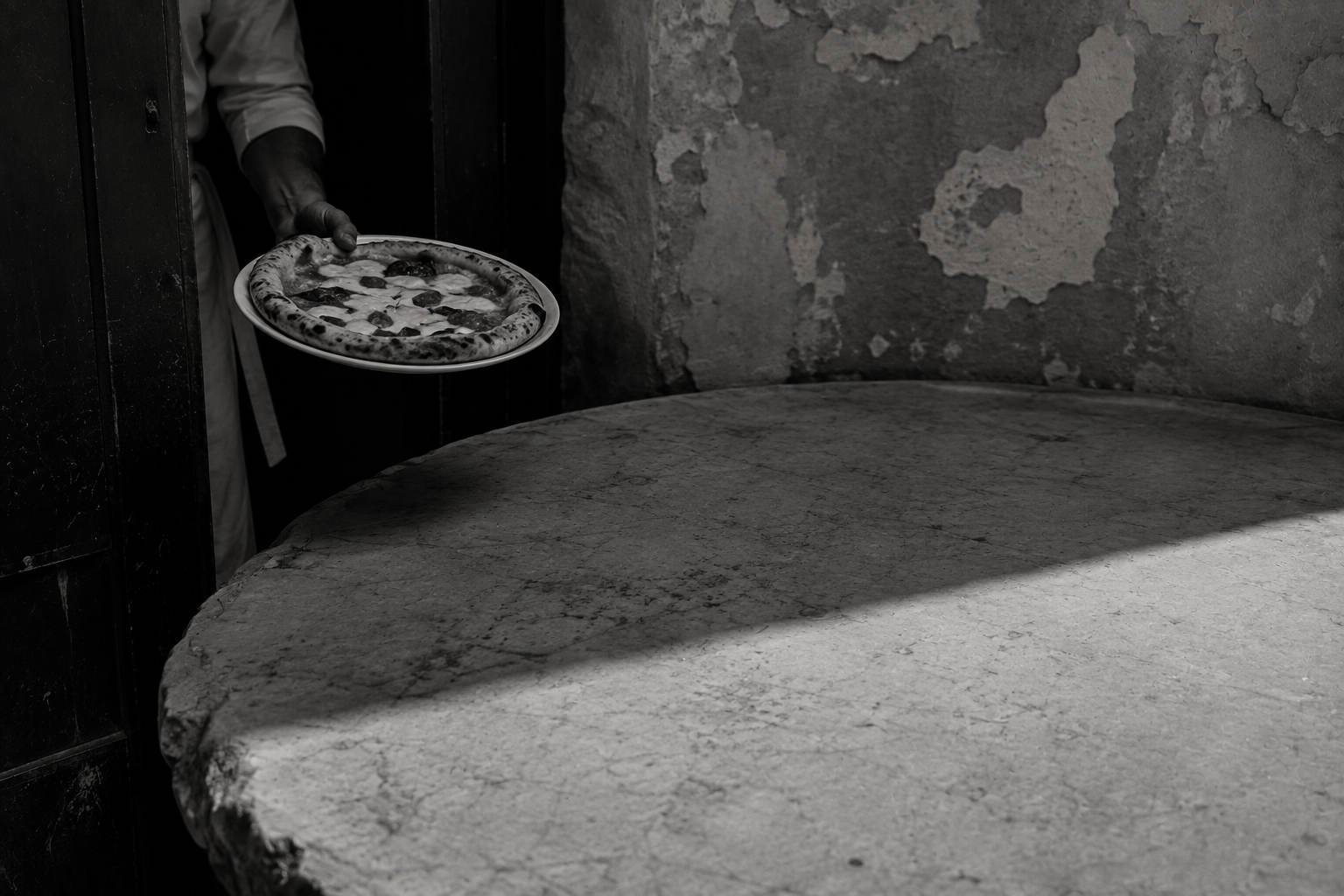 Black-and-white editorial image of a person carrying a pizza toward a large weathered stone table, with dramatic sunlight and deep architectural shadows.