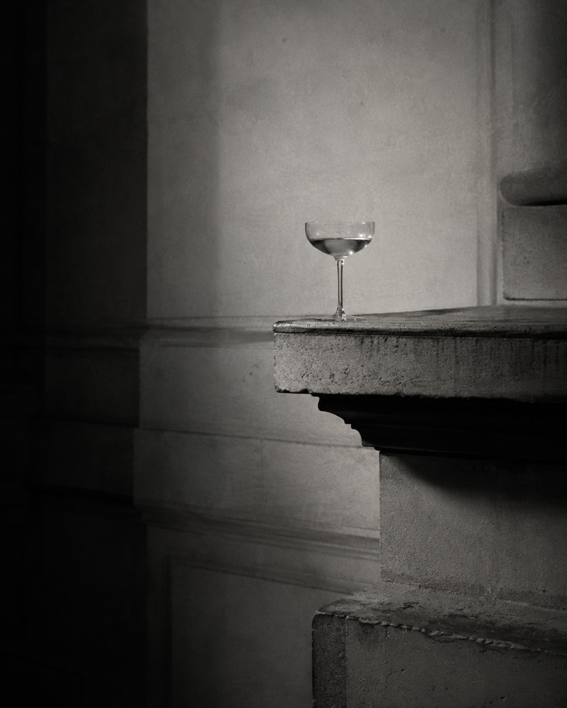 A black and white photo of a cocktail glass with a long stem, placed on a concrete ledge in a minimalist setting.