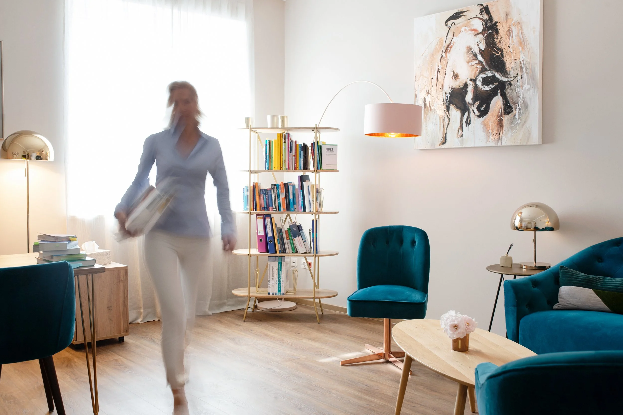 Ein modernes Wohnzimmer mit blauen Sesseln, einem Bücherregal, einem abstrakten Wandbild und einer Person, die durch den Raum geht, während sie ein Buch hält.