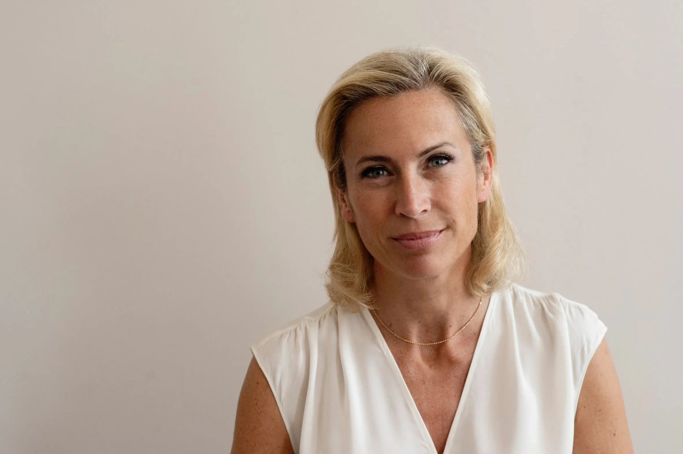Naheaufnahme einer lächelnden, blonden Frau im weißen Kleid gegen eine helle Wand.