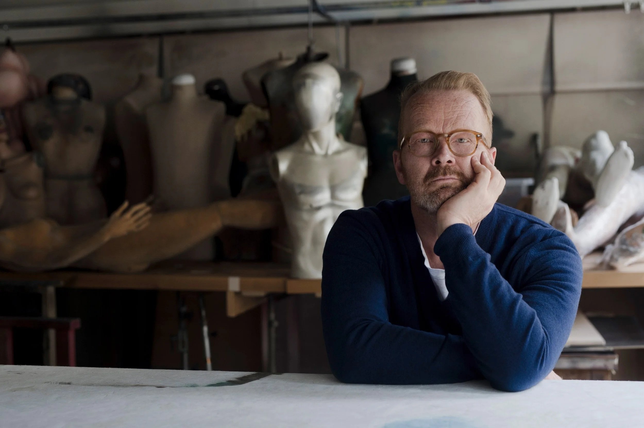 Ein Mann sitzt nachdenklich an einem Tisch in einem Raum mit Kunstskulpturen und -mannequins im Hintergrund.