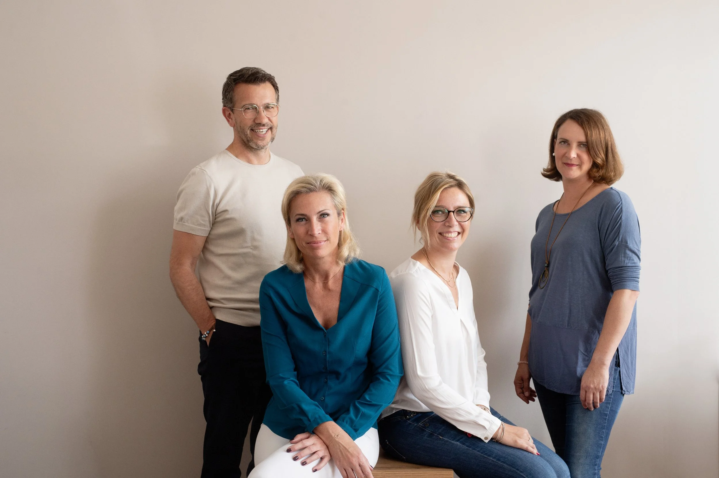 Fünf Erwachsene, drei Frauen und zwei Männer, posieren vor einer weißen Wand. Zwei Frauen sitzen, die anderen stehen. Sie sehen freundlich und professionell aus.