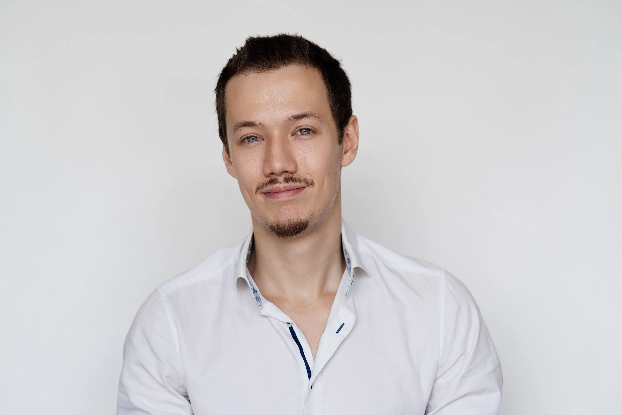 Porträt eines Mannes mit dunklen kurzen Haaren, blauen Augen, leichten Stirnfalten, einem kleinen Kinnbart und einem freundlichen Lächeln, getragen in einem weißen Hemd vor neutralem hellgrauem Hintergrund.