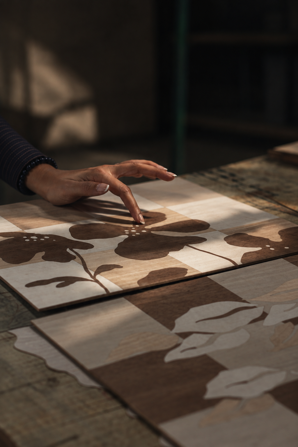 A person's hand is touching a wooden art piece featuring of a botanical design with leaves and stems, arranged on a table with other similar wood art pieces.