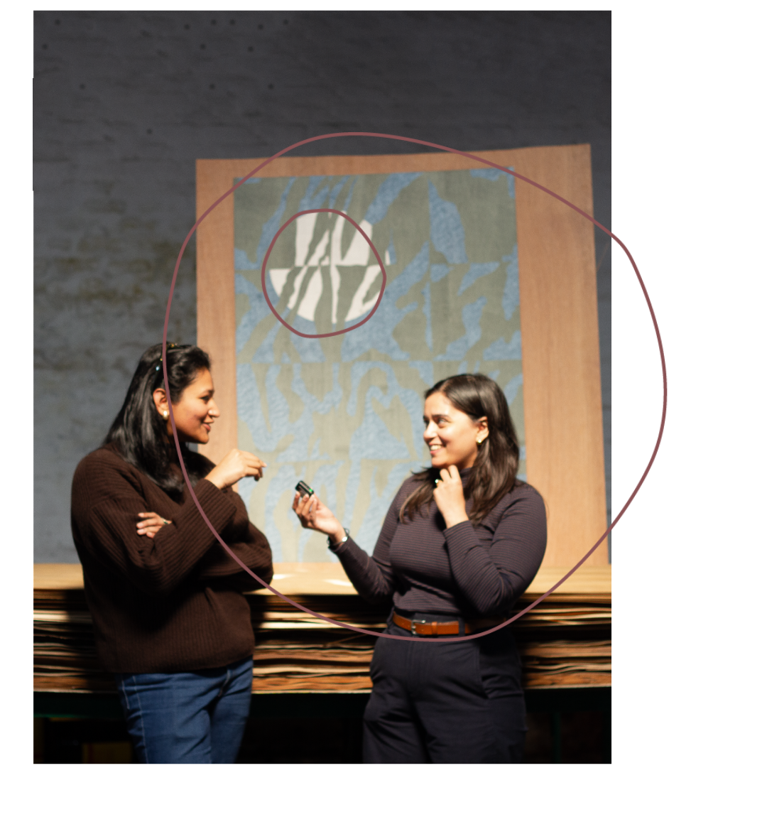 Two women are standing and talking in a room with a textured wall and large artwork in the background. One woman is holding a small object, and they are smiling at each other.