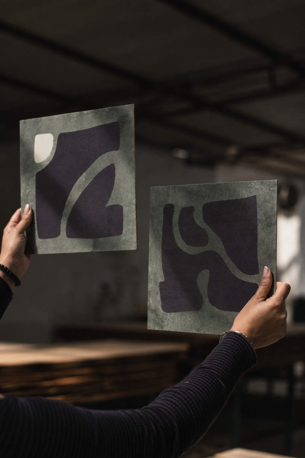 Person holding abstract art tiles with dark shapes and greenish backgrounds in a workshop or studio.