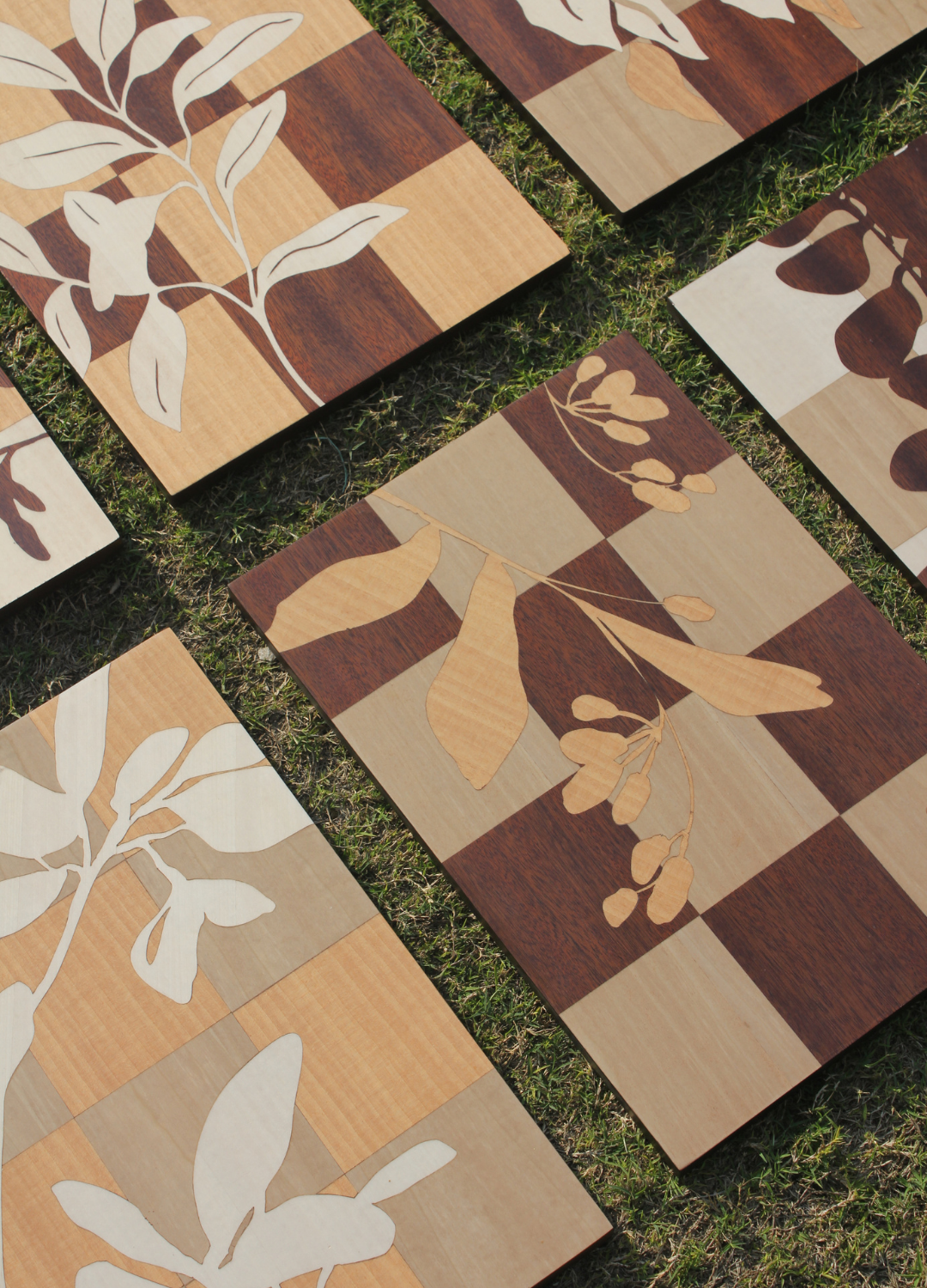 Multiple wooden chessboards with leaf and botanical patterns, placed on grass.