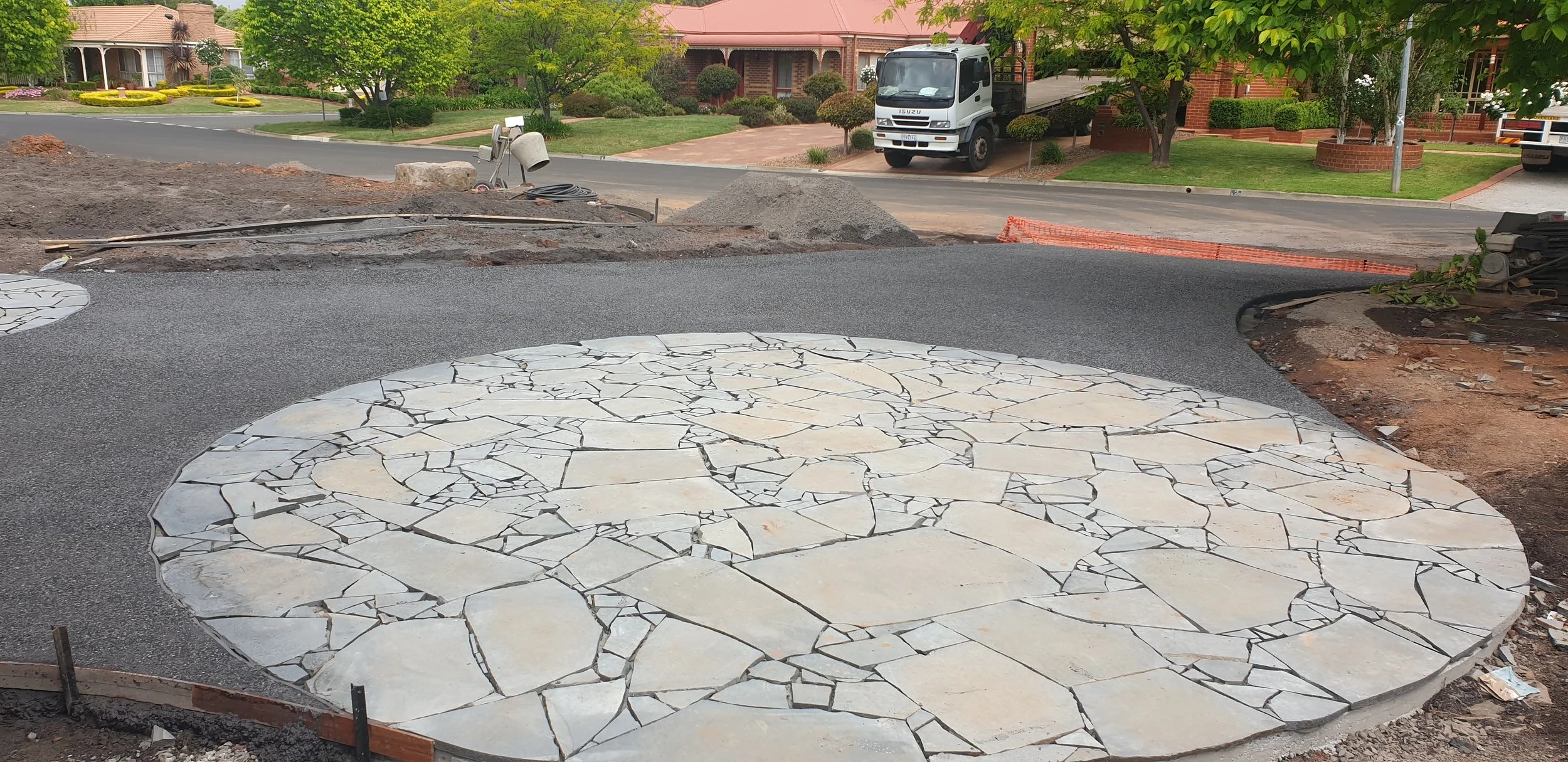 Construction site with a circular stone pavement area in the foreground and ongoing asphalt work behind it, with construction tools and materials nearby.