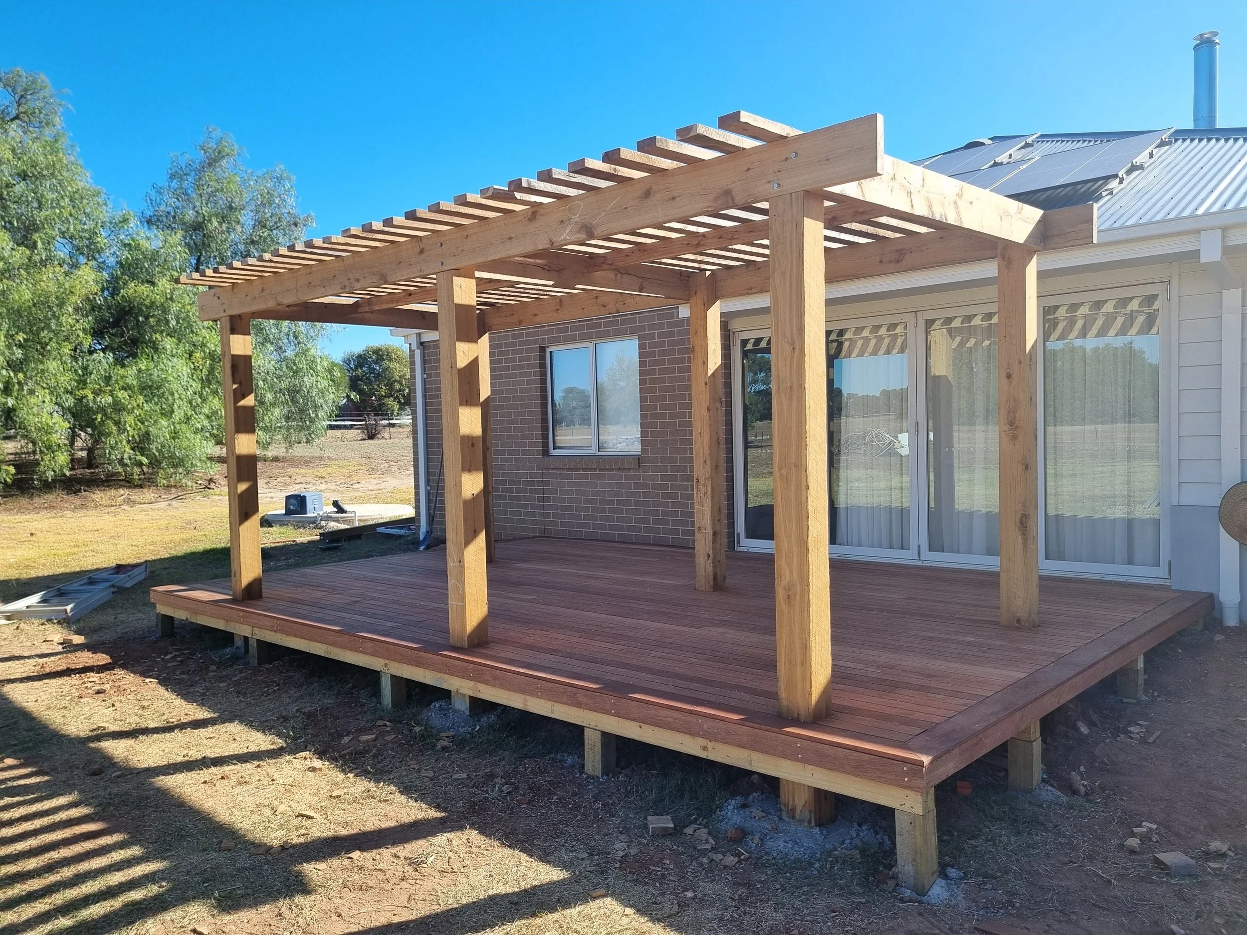 A wooden deck structure under construction attached to the back of a house, with a pergola framework on top, in a backyard with trees and a clear blue sky.