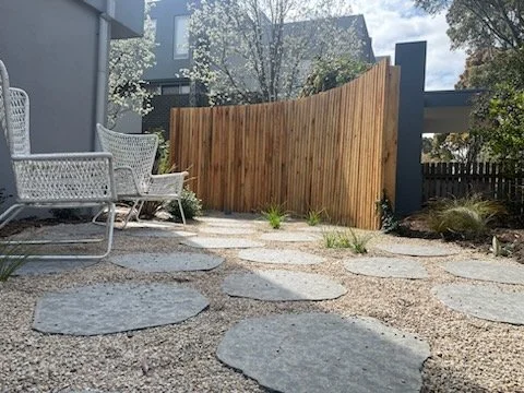 A backyard patio area with two white wicker chairs, a wooden privacy fence, stone pathway, and some plants.