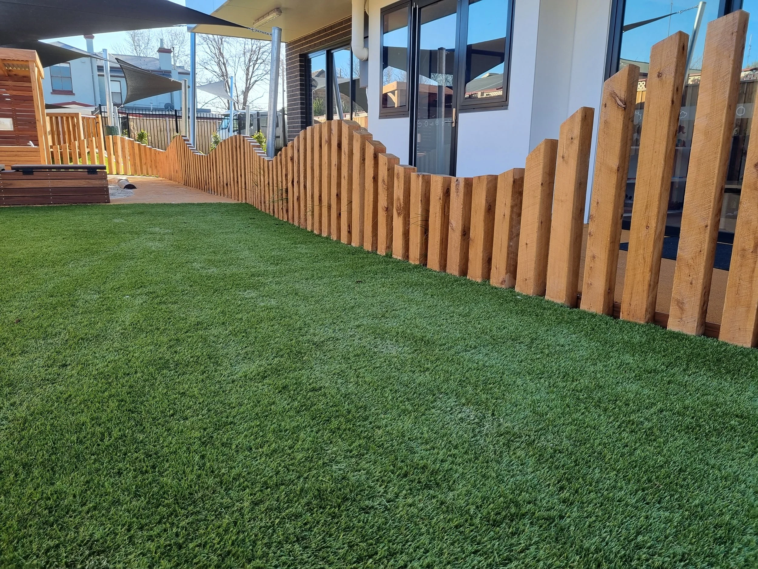 View of a backyard with a wooden fence, artificial grass, and a house with large glass windows and sliding doors.