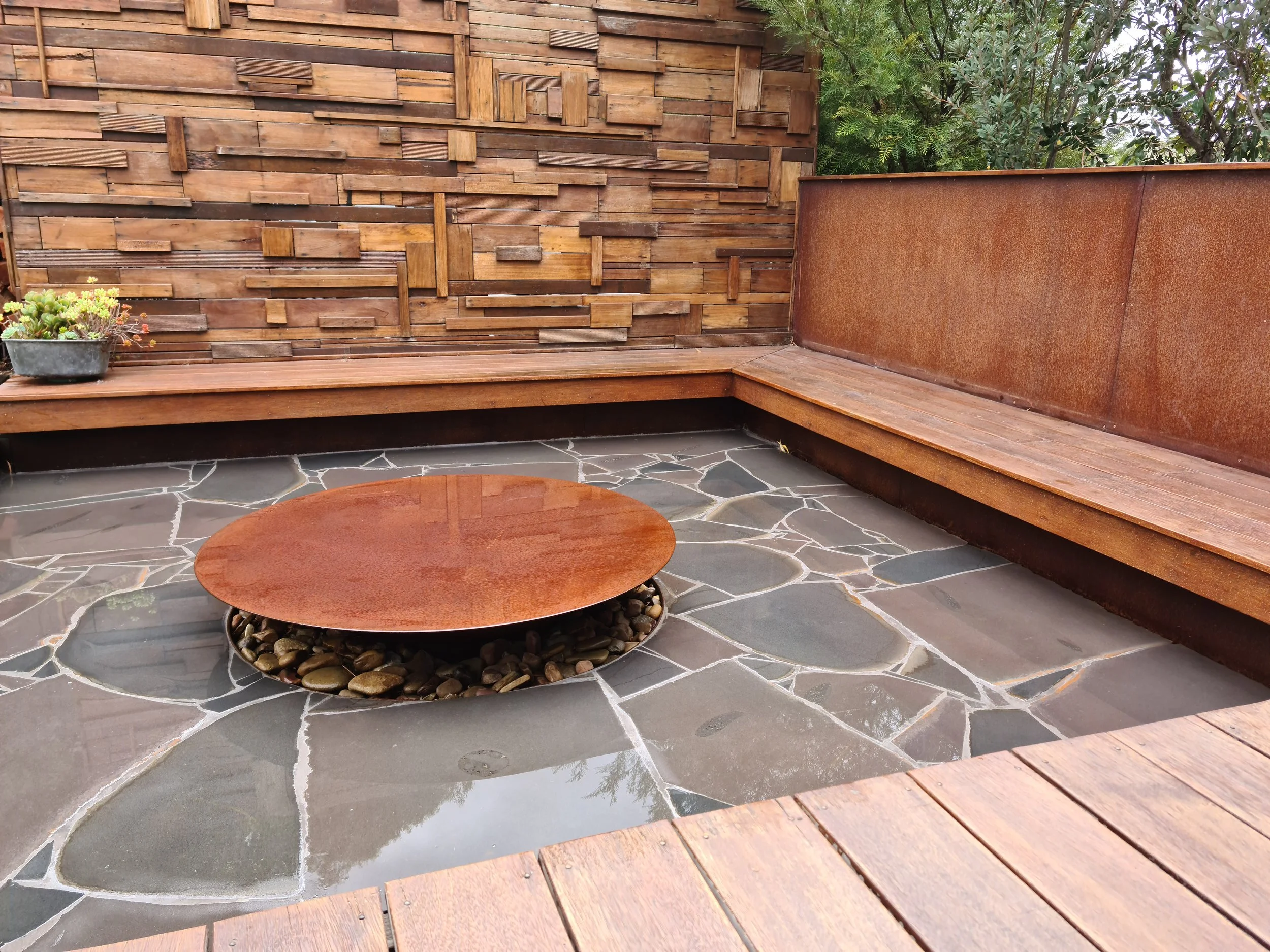 An outdoor corner patio with a wood slat wall, wooden built-in bench, rust-colored metal side wall, potted plant with succulents, and a stone mosaic patio surface with a circular fire pit filled with smooth rocks.