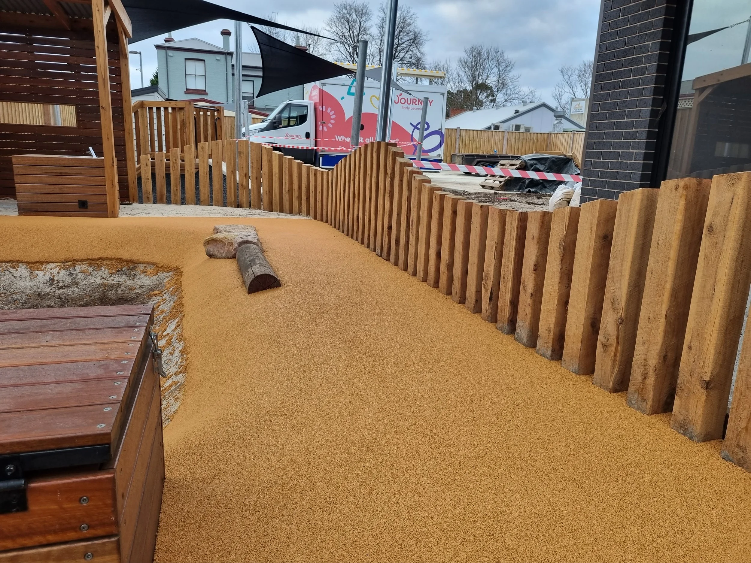 A fenced outdoor play area with wood and rubber flooring, a wooden bench, and construction equipment in the background including a truck and building materials.