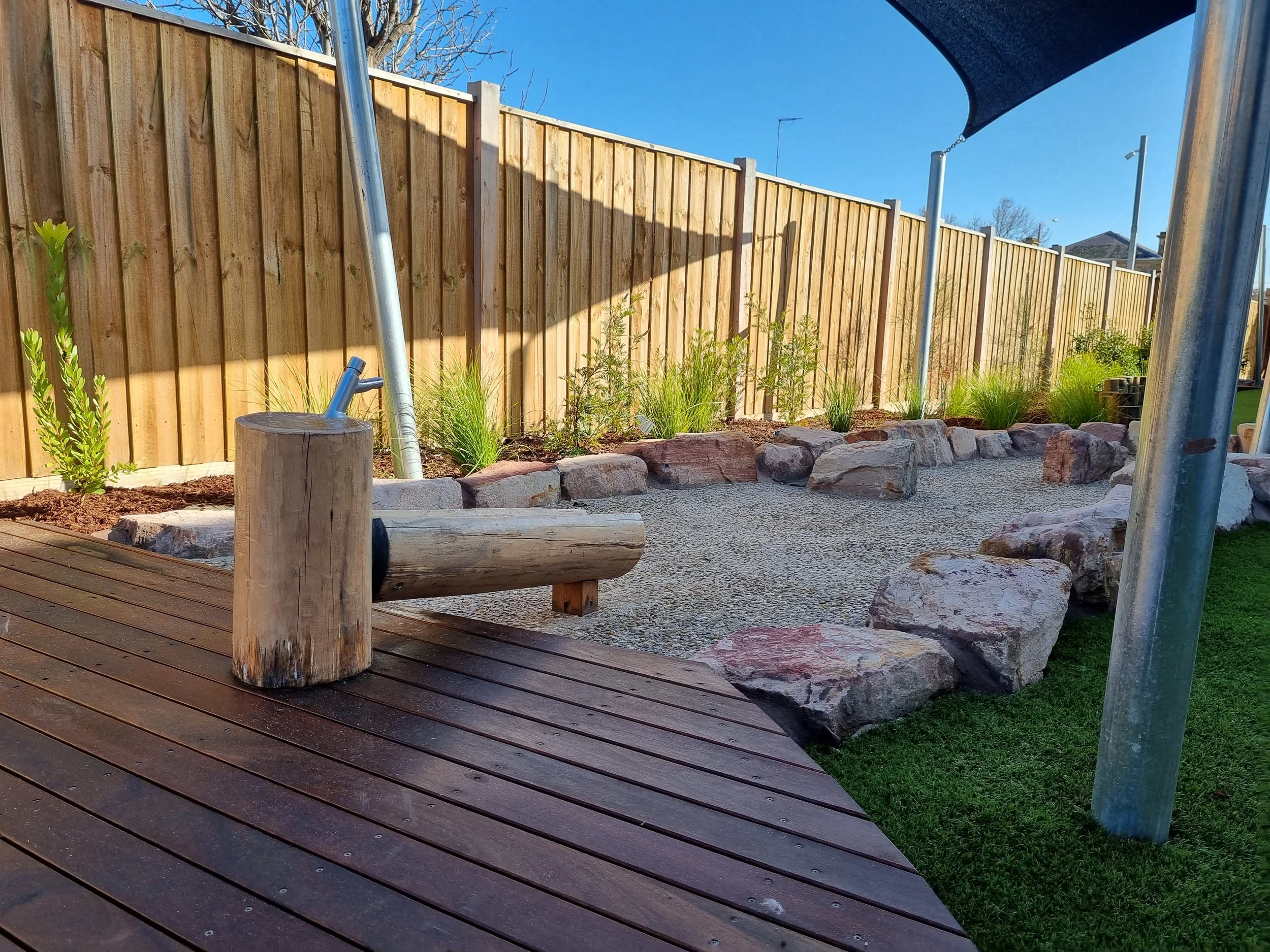 A backyard featuring a wooden privacy fence, a small garden bed with green plants and rocks, a wooden stepping log, a gravel pathway, and part of a trampoline with a shade cover, with clear blue sky above.