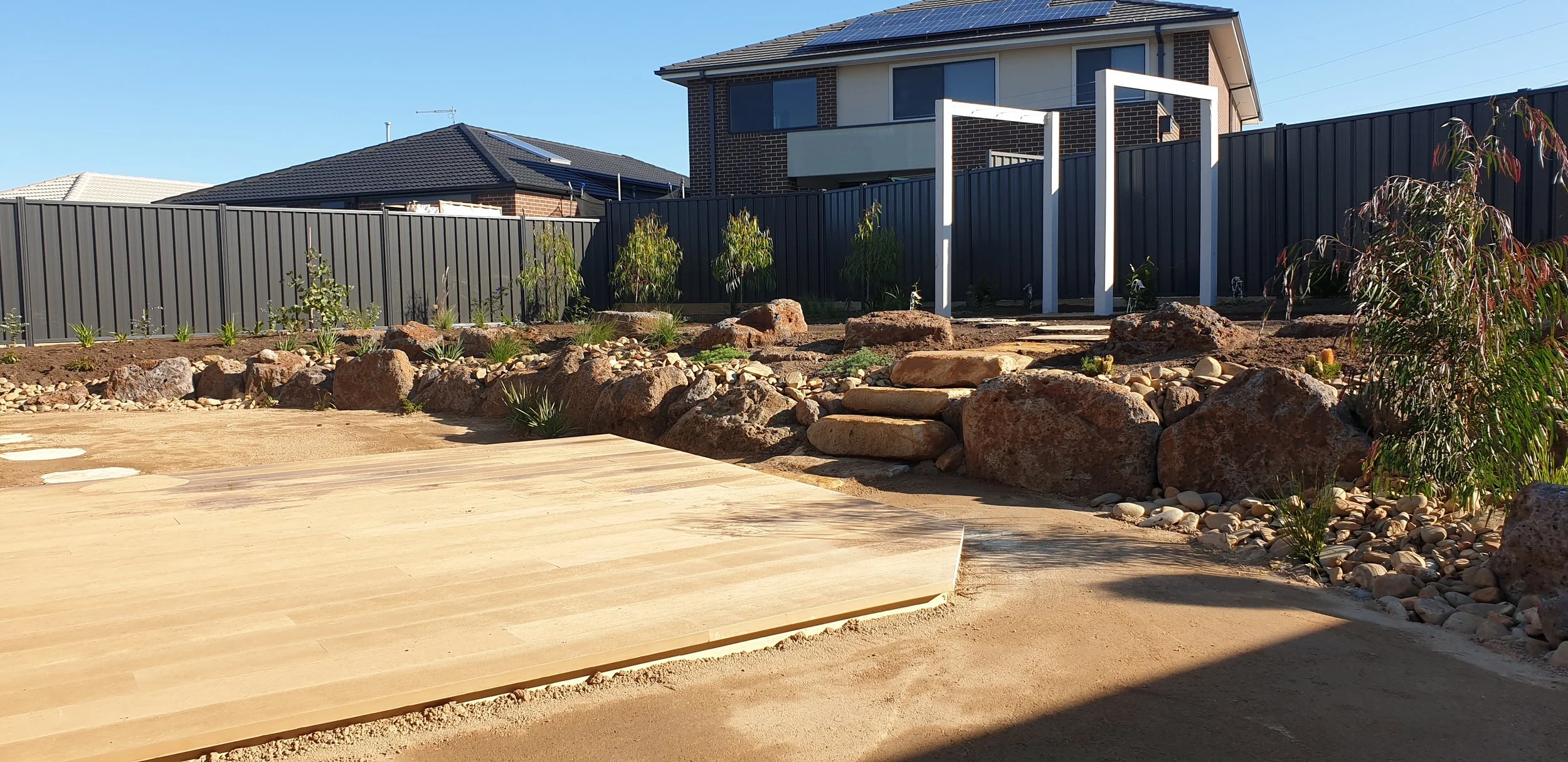 A residential backyard with a climbing frame structure, landscaped with rocks, plants, and a wooden deck, with houses and solar panels in the background.