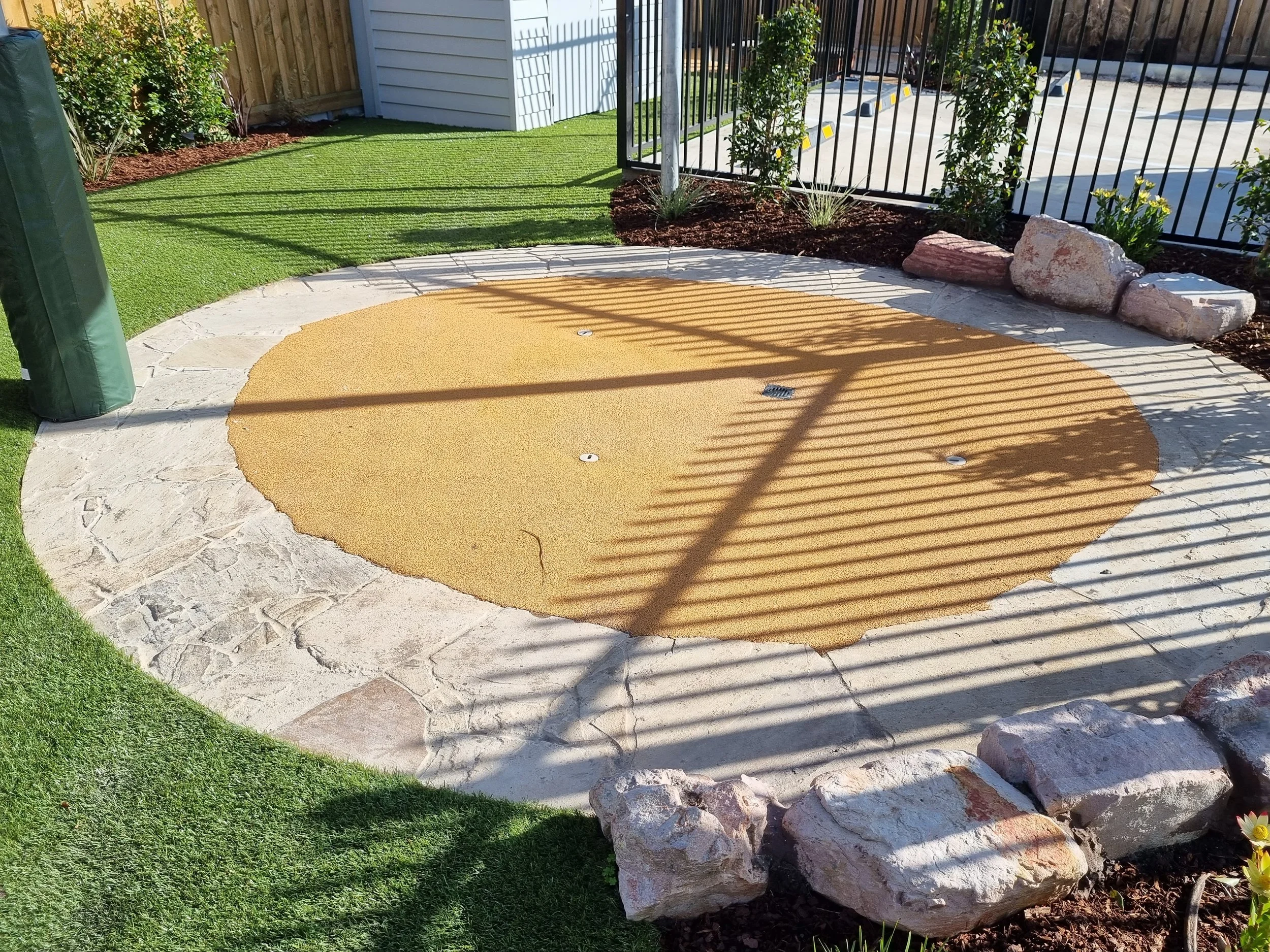 A landscaped backyard with artificial grass, a concrete patio, flowering plants, and decorative rocks, enclosed by a metal fence and wooden fence.