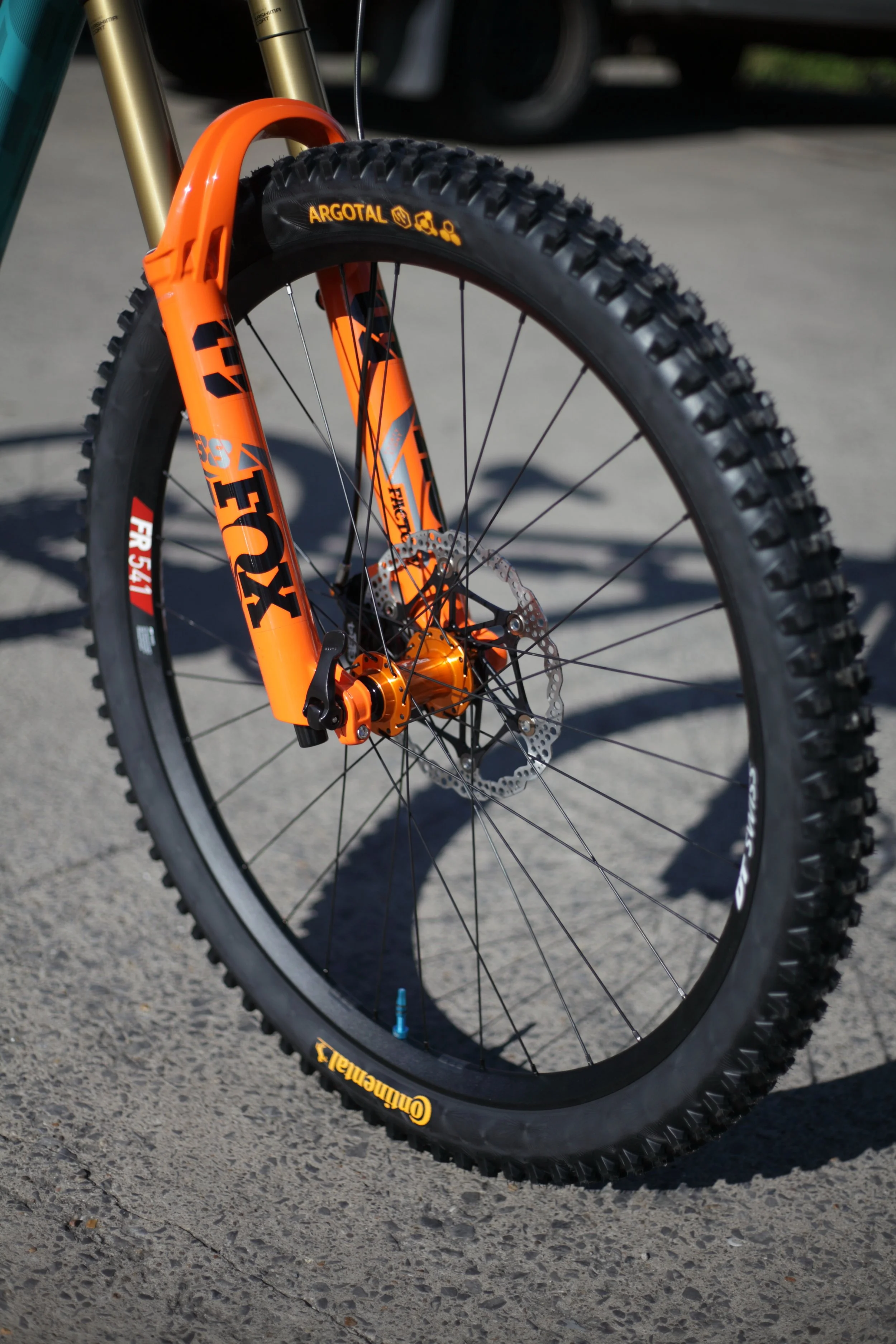 Close-up of an orange mountain bike front wheel with high-performance tires, disc brake rotor, and suspension fork, parked on a paved surface.