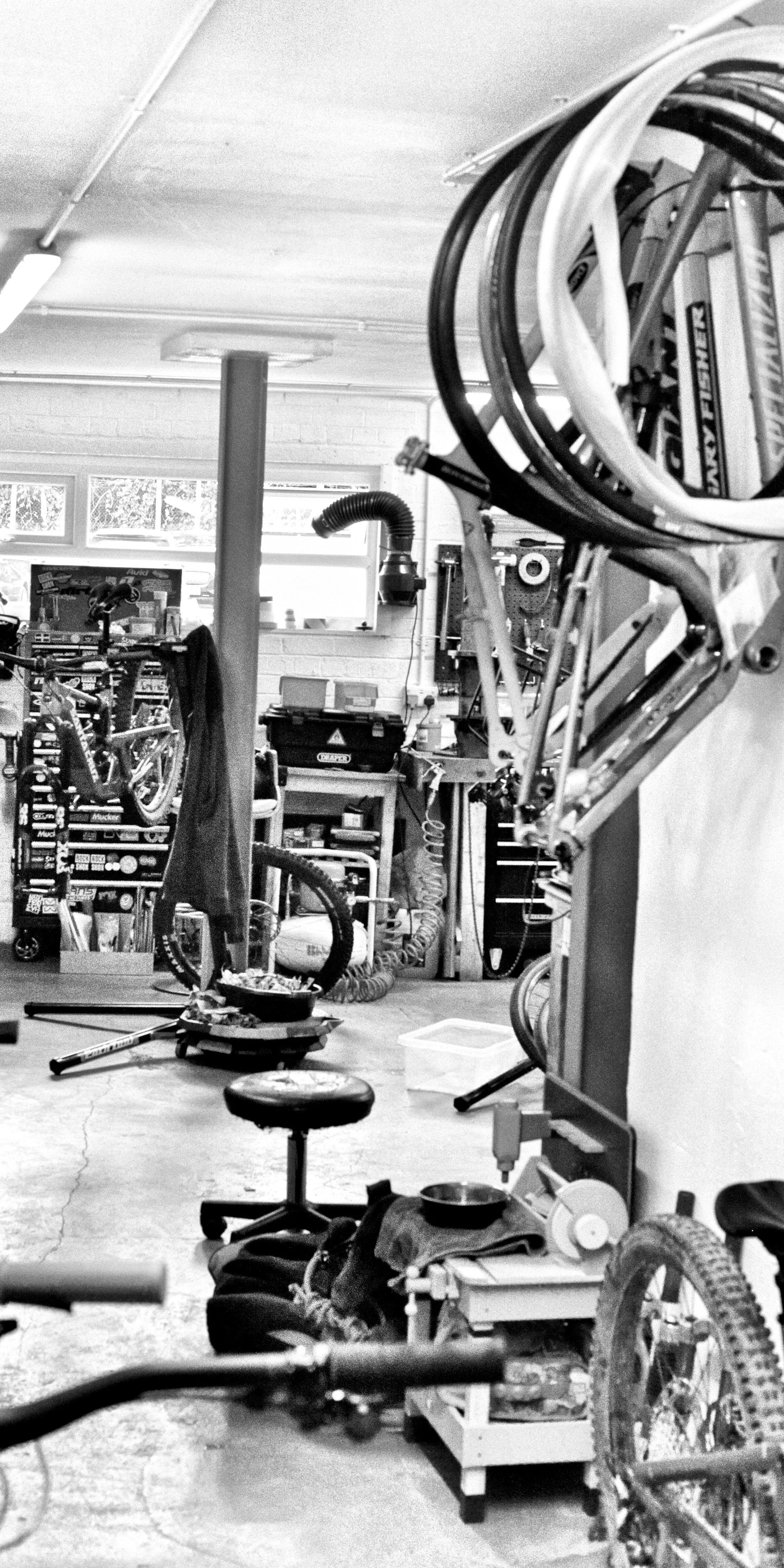 A cluttered bike repair workshop with bikes hanging from the ceiling and scattered tools and equipment on the floor and workbenches.