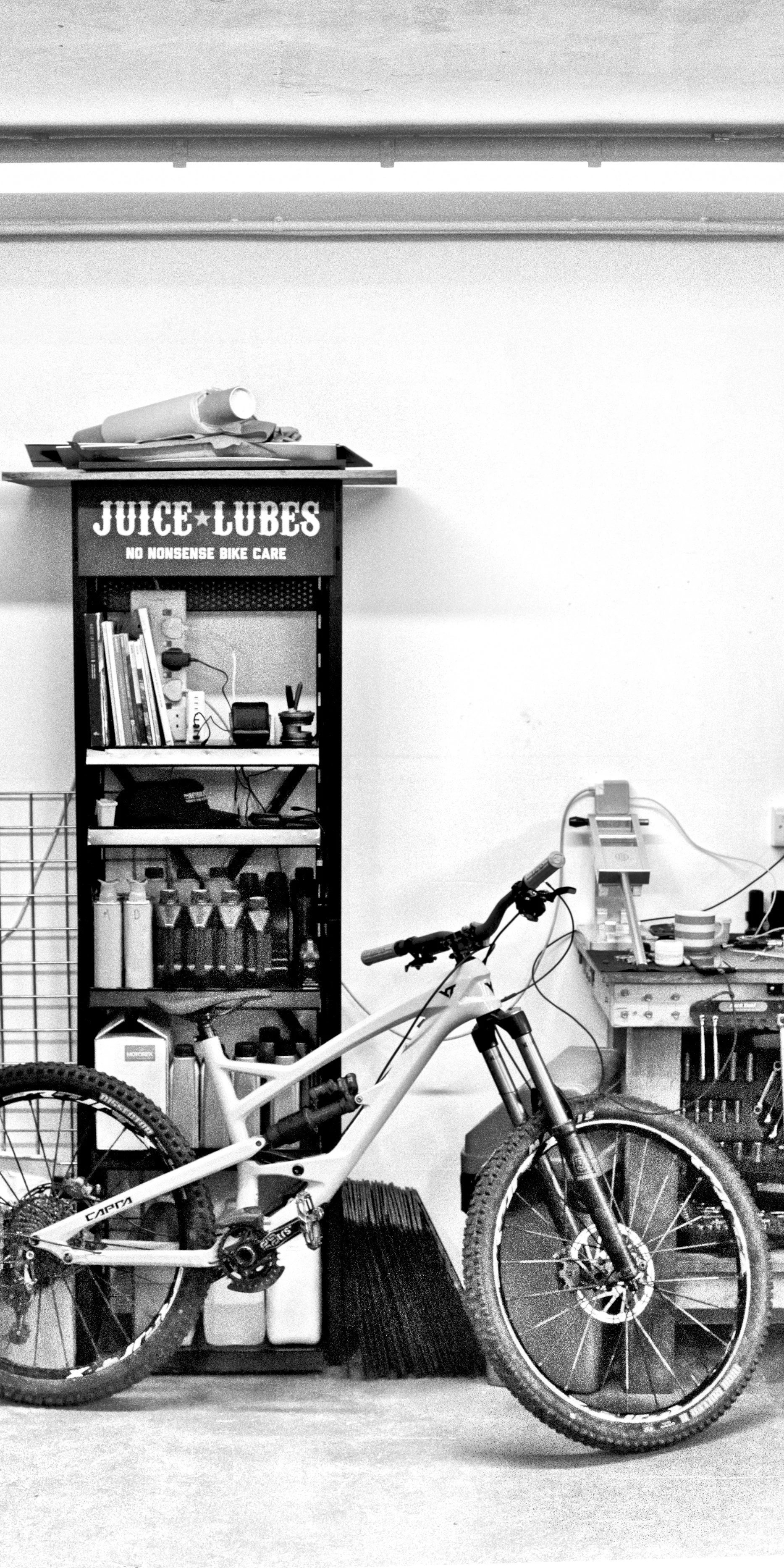 Indoor scene with a mountain bike in front of a black metal shelf filled with bottles, cans, and books. There is a sign on the shelf that reads 'Juice Lubes No Nonsense Bike Care.' Various tools and supplies are on a workbench to the right, with wires and small containers. A coffee cup and a paper towel roll are on the top of the shelf, and part of a wire grid is visible on the far left.