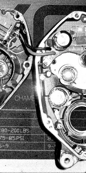 Close-up of an internal engine component with gears, bolts, and wiring, shown in black and white.