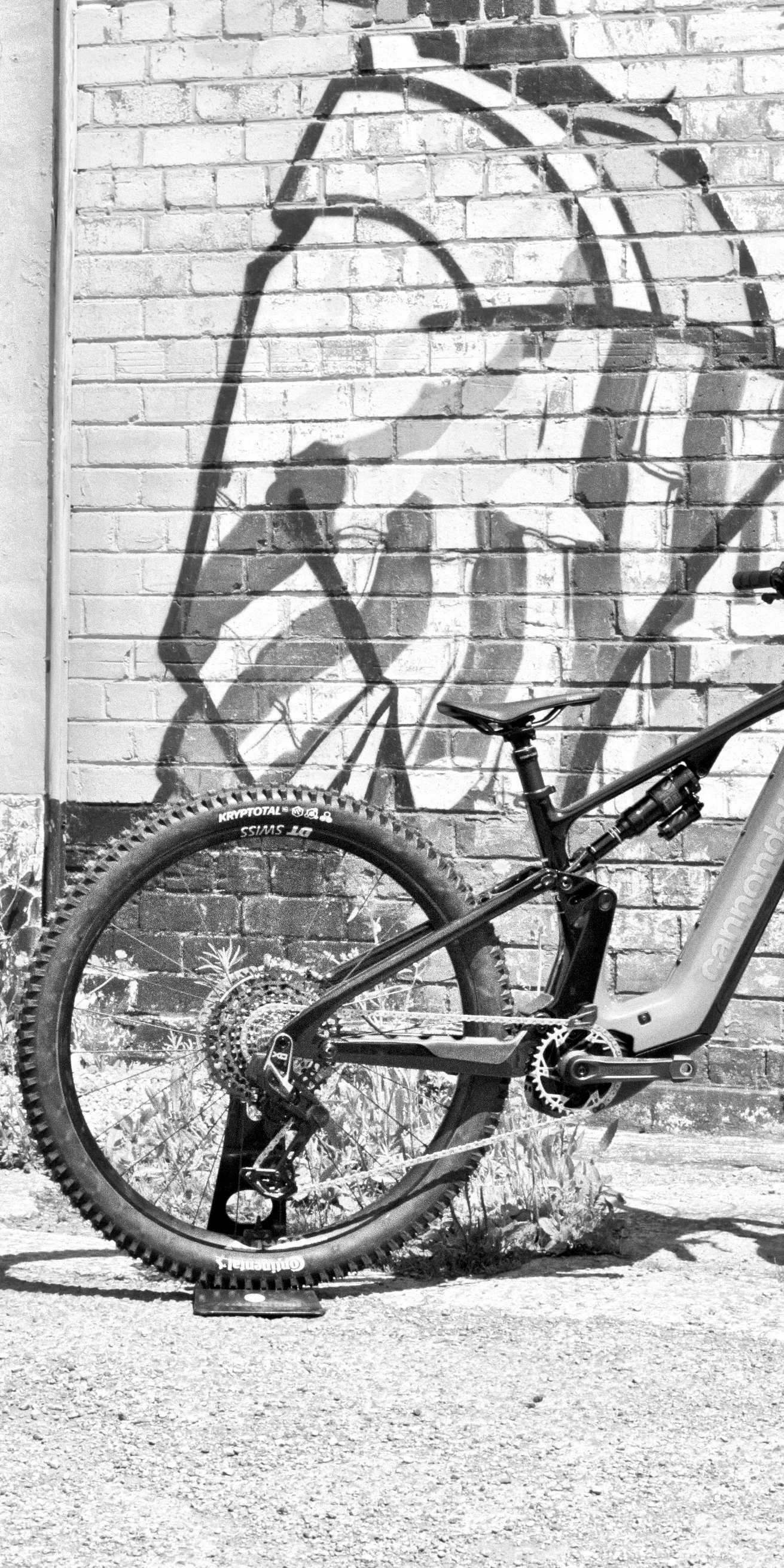 A mountain bike with thick, knobby tires and advanced suspension system, leaning against a brick wall with a large street art mural of a stylized hand holding a spray paint can.