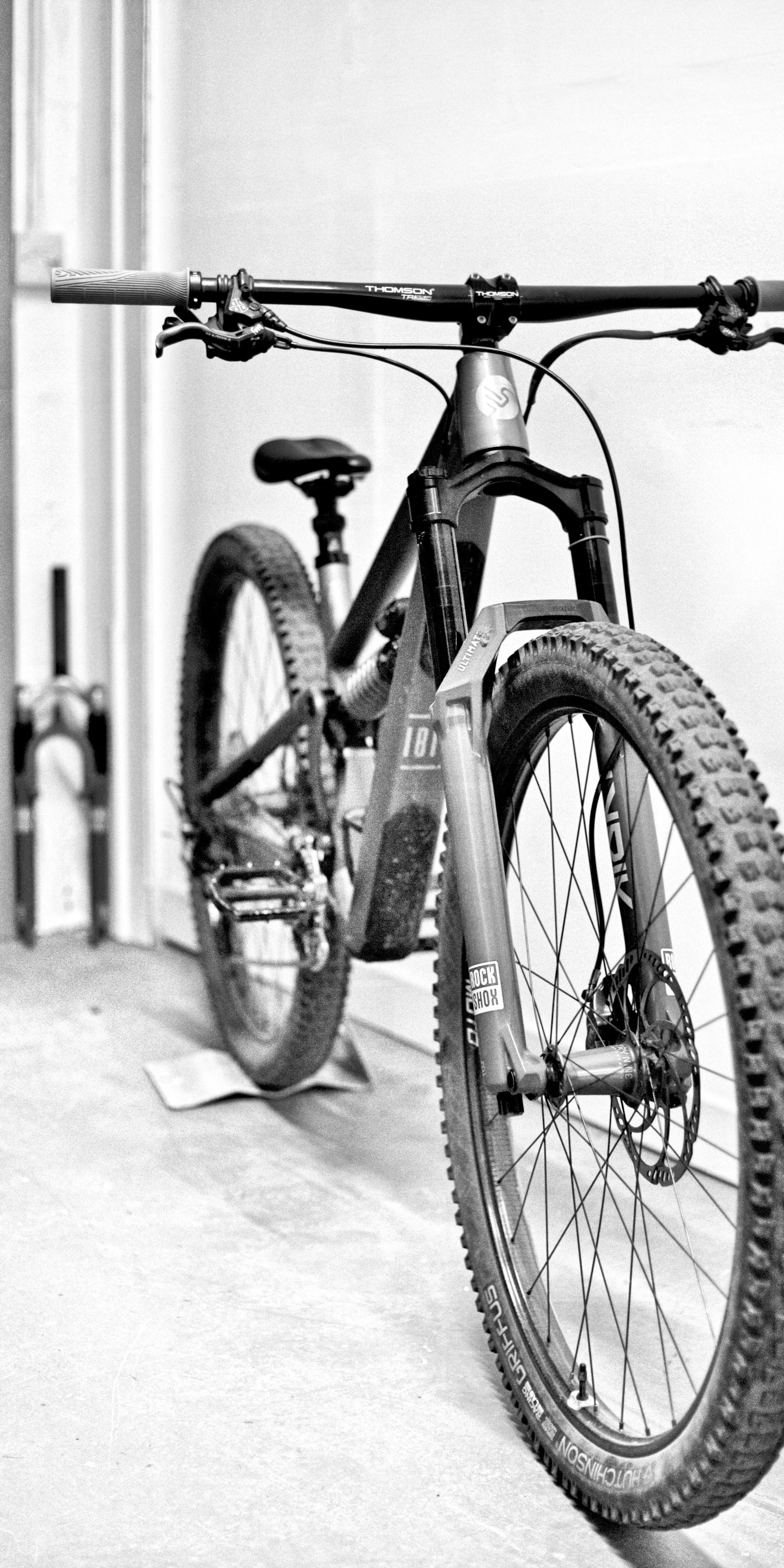 A black and white photograph of a mountain bike, showing the front wheel, handlebars, and frame, positioned indoors against a plain wall.