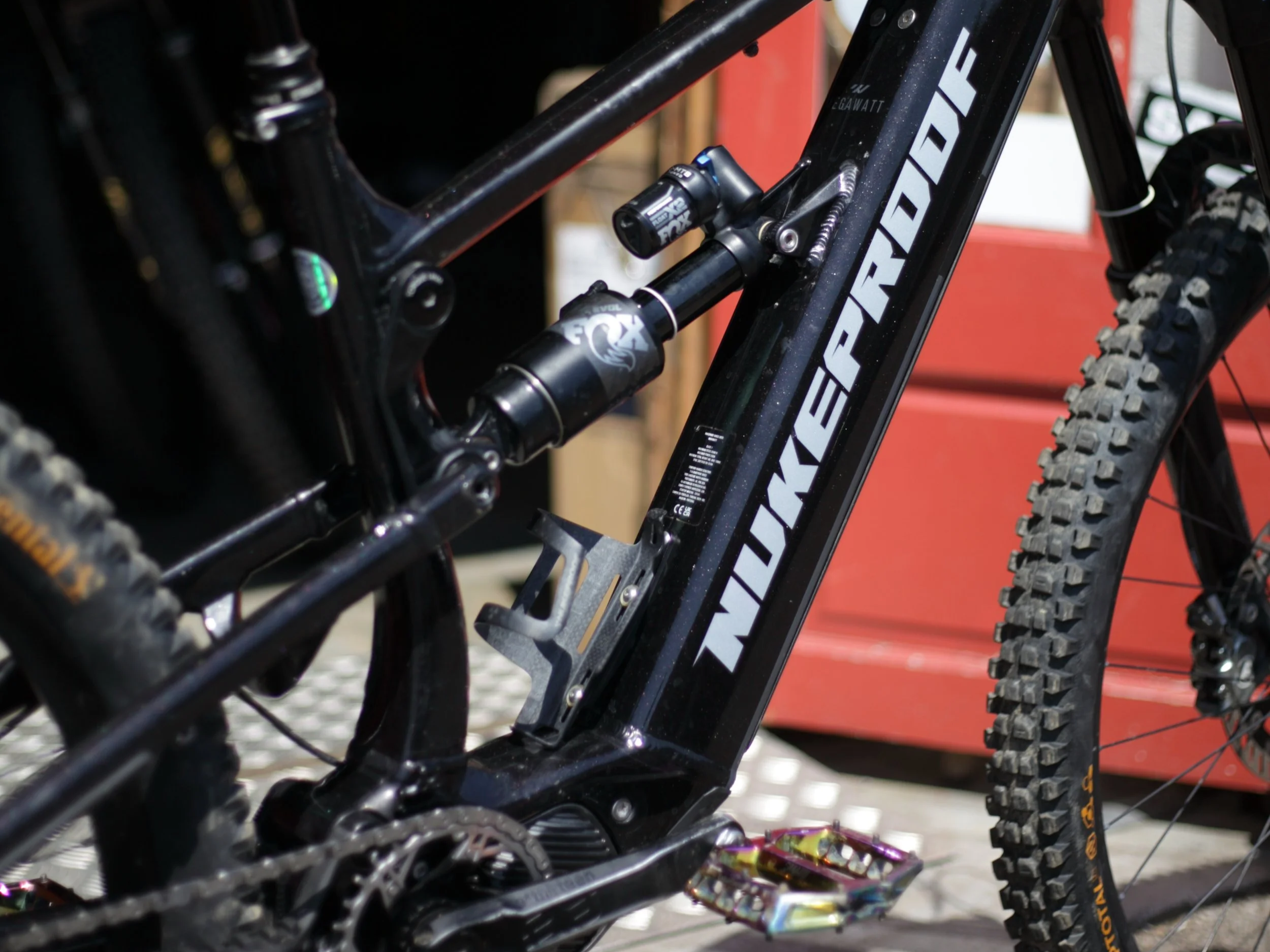 Close-up of a black mountain bike frame with Fox suspension, large knobby tires, and a pedal, with colorful wrapped candies and a red toolbox in the background.