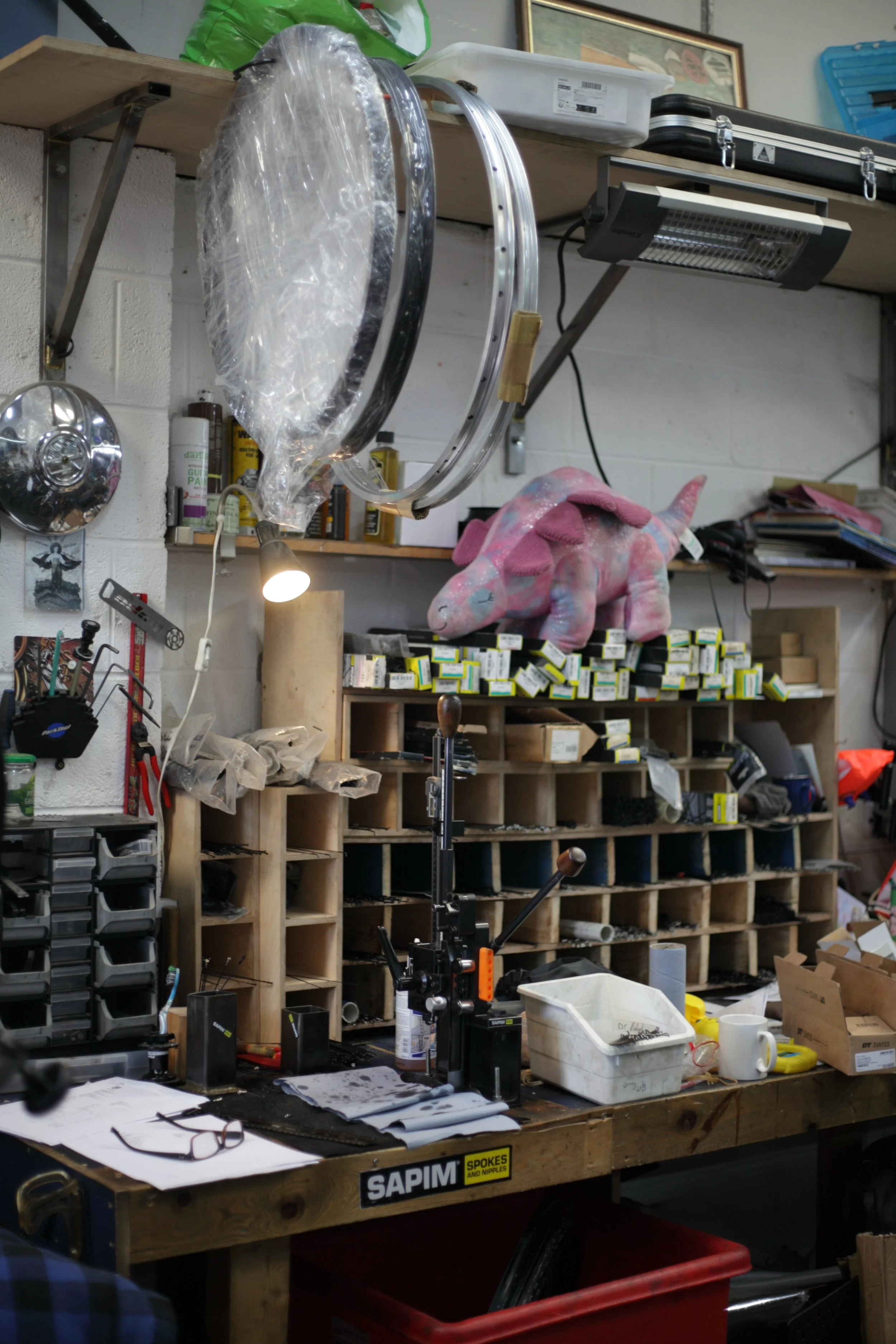 A cluttered workshop or workbench with various tools, parts, and supplies. There is a pink stuffed animal on a shelf, and a large circular object wrapped in plastic hanging from the ceiling. Several shelves filled with small containers and boxes are visible in the background.