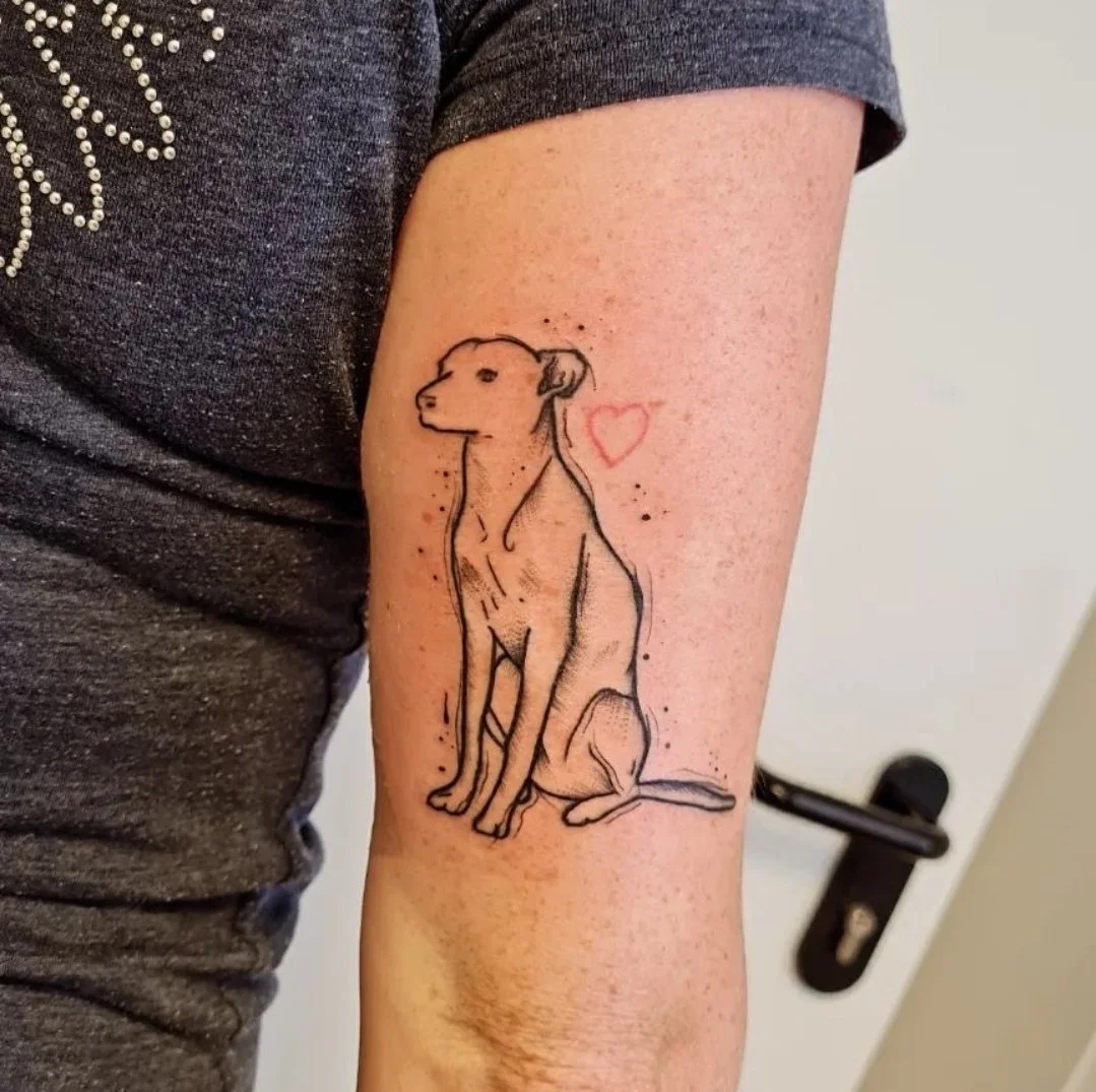 Tattoo eines sitzenden Hundes mit einem kleinen roten Herz daneben auf einem Arm.