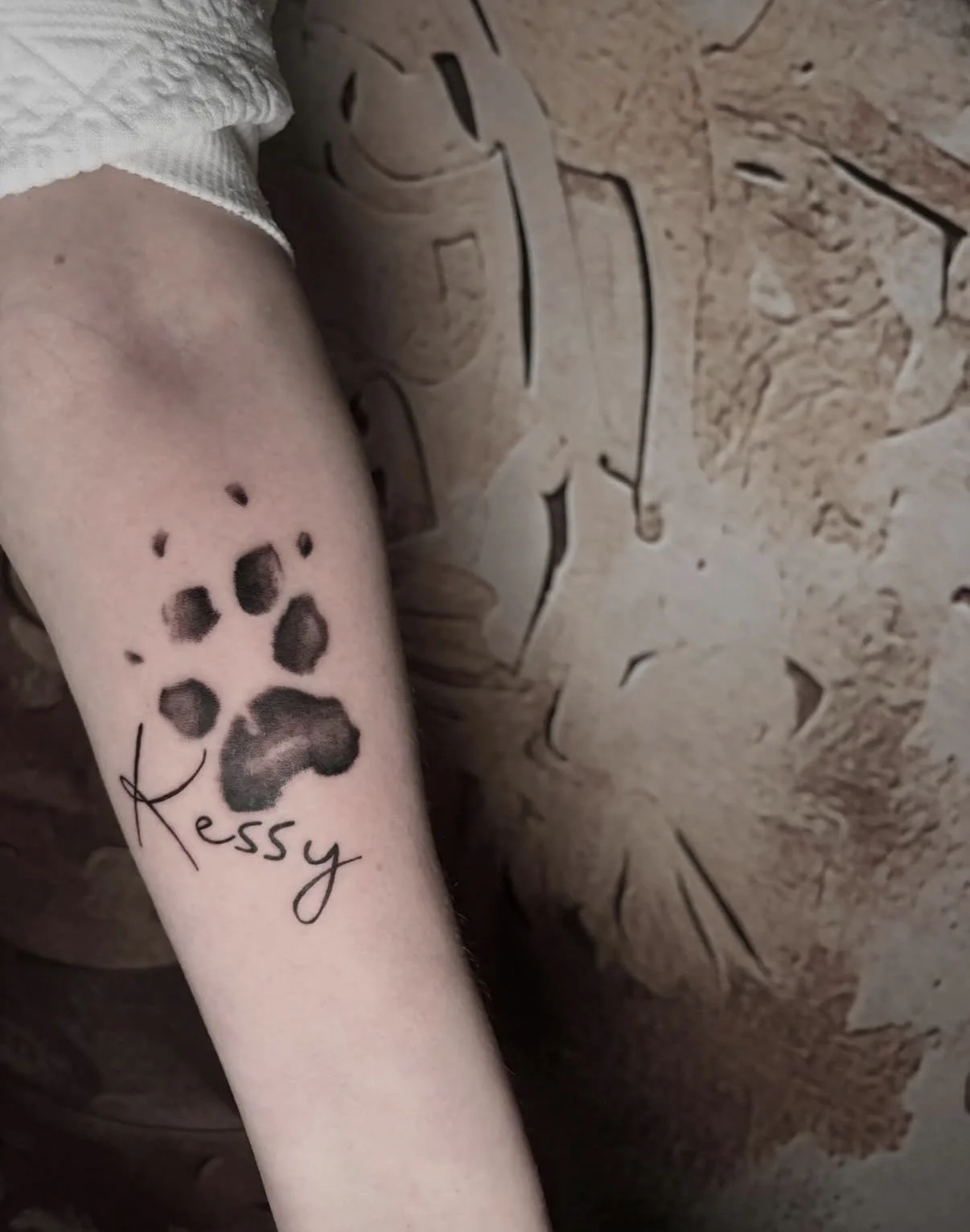 Tattoo eines Hundepfotenabdrucks mit dem Namen 'Kessy' auf dem Arm, vor einem Steinmauer-Hintergrund.
