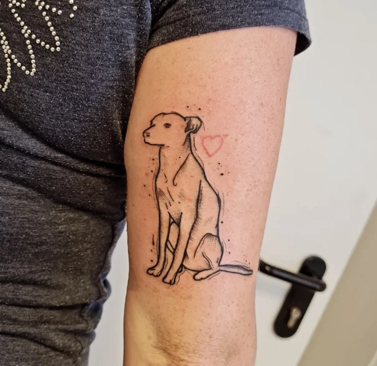 Tattoo eines sitzenden Hundes mit Herz daneben auf den Arm einer Person.