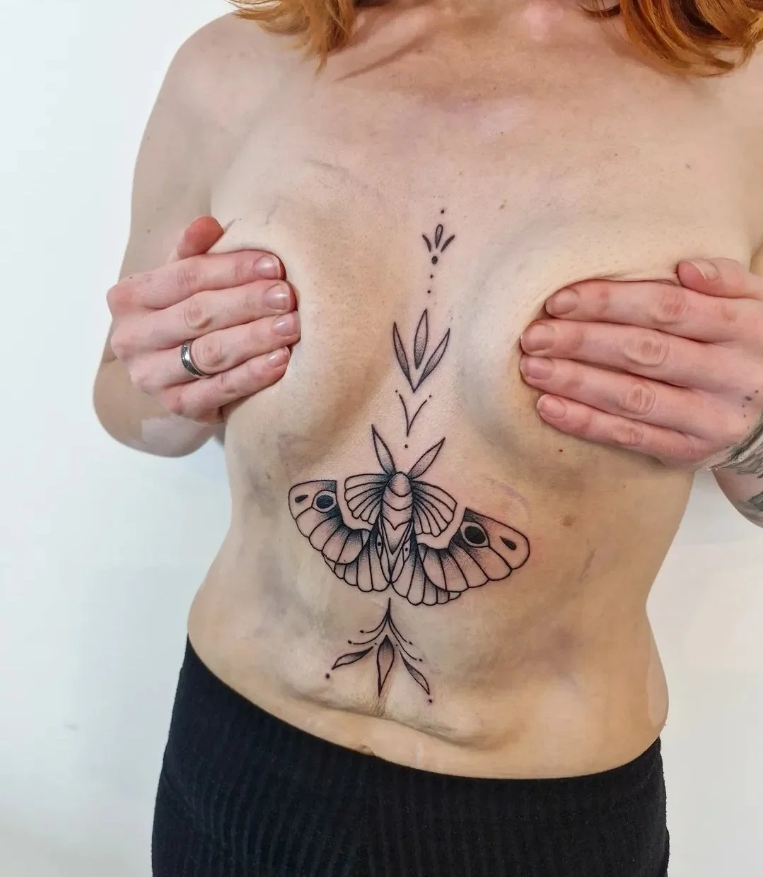 Eine Frau mit tätowiertem Schmetterling auf dem Bauch, die ihre Brüste mit den Händen bedeckt.