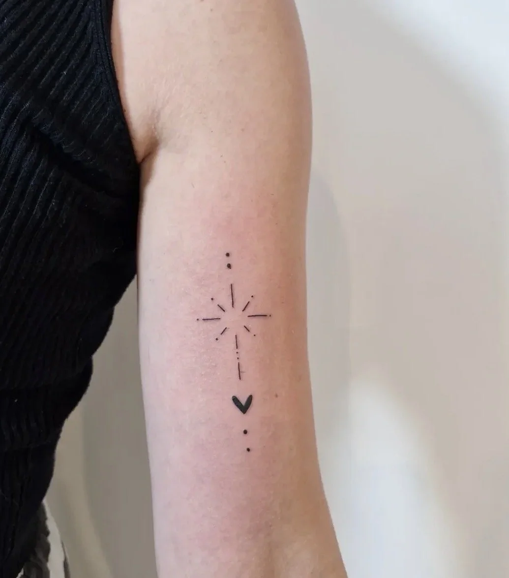 Tattoo auf dem Oberarm mit einem Kreuz und einem Herz, umgeben von Punkten und Linien.