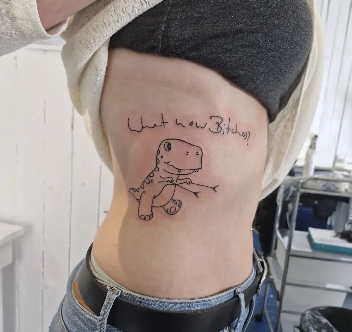Eine Person mit einer Tattoo-Zeichnung auf dem Rücken. Das Tattoo zeigt einen lustigen Dinosaurier mit einem freundlichen Gesicht und auch den Text "What how Bitches" darüber.